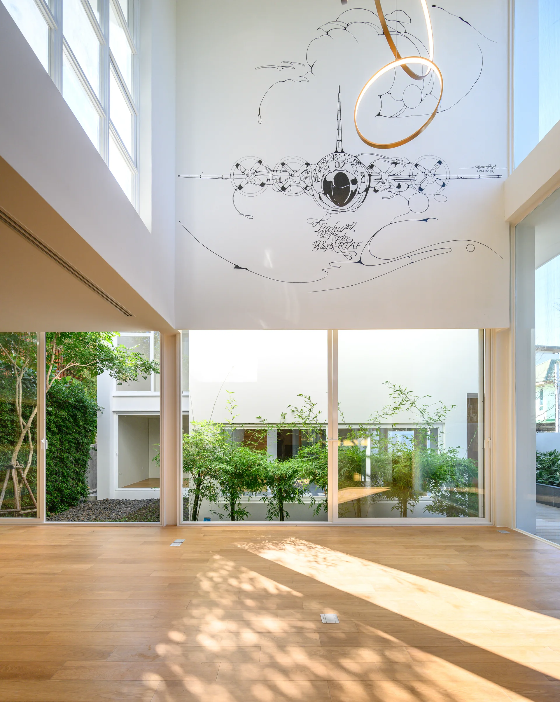 A bright double-height room features light wood floors, large glass windows viewing a courtyard, and a black line art airplane mural on a high white wall.