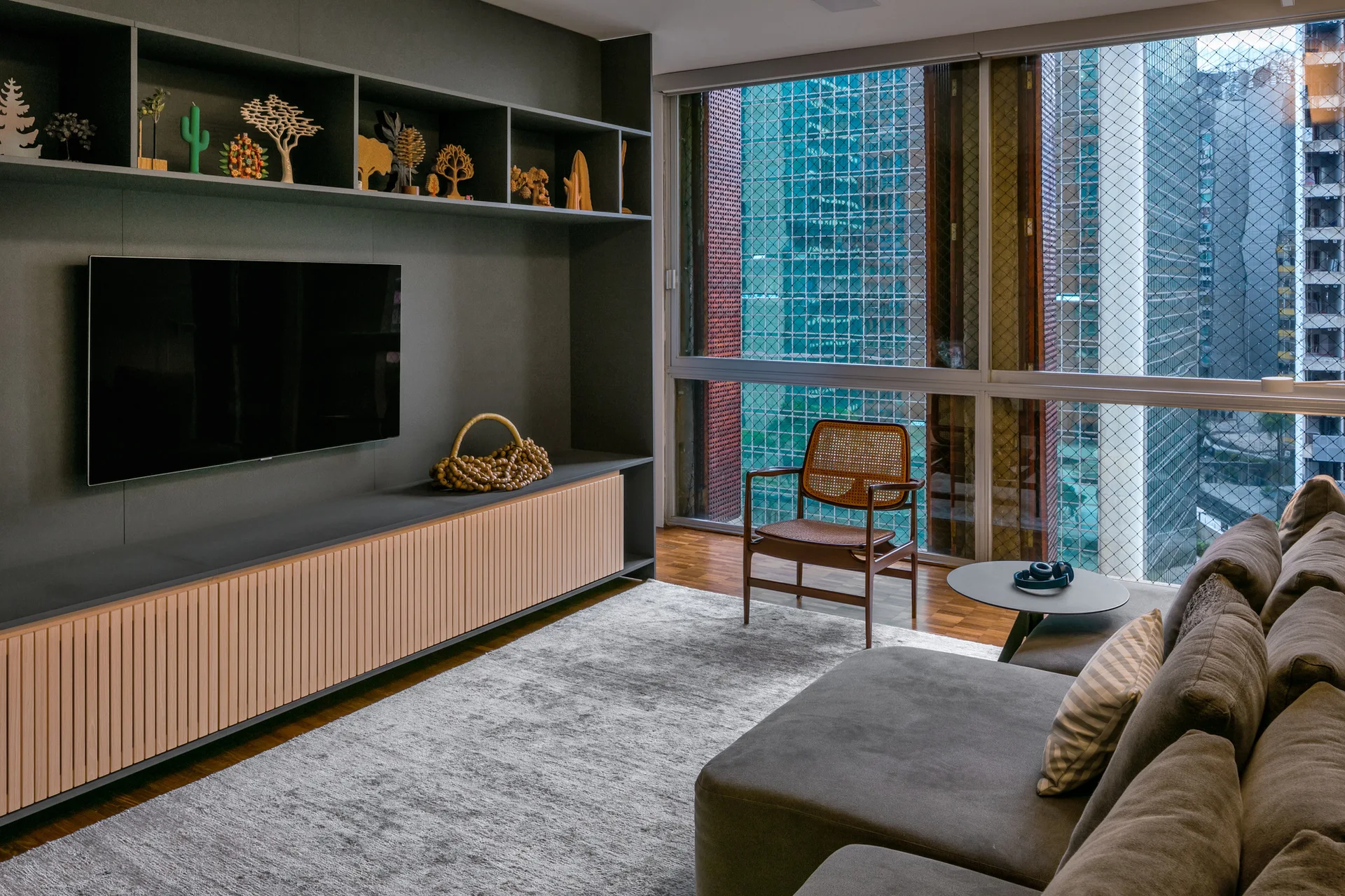 A modern grey living room featuring a matte charcoal built-in media wall, ribbed wood cabinets, and a cane armchair near large windows.