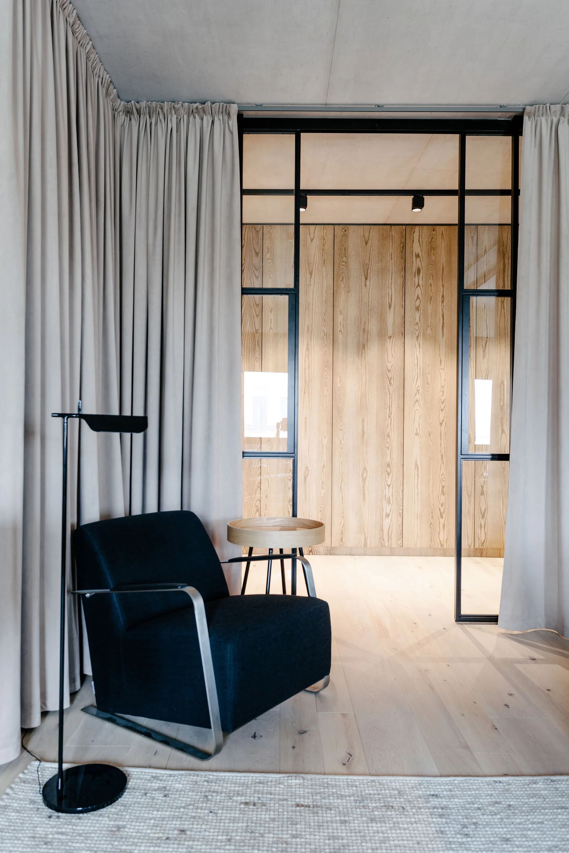A black fabric armchair and thin floor lamp stand in front of beige drapes next to a black steel-framed glass partition door.