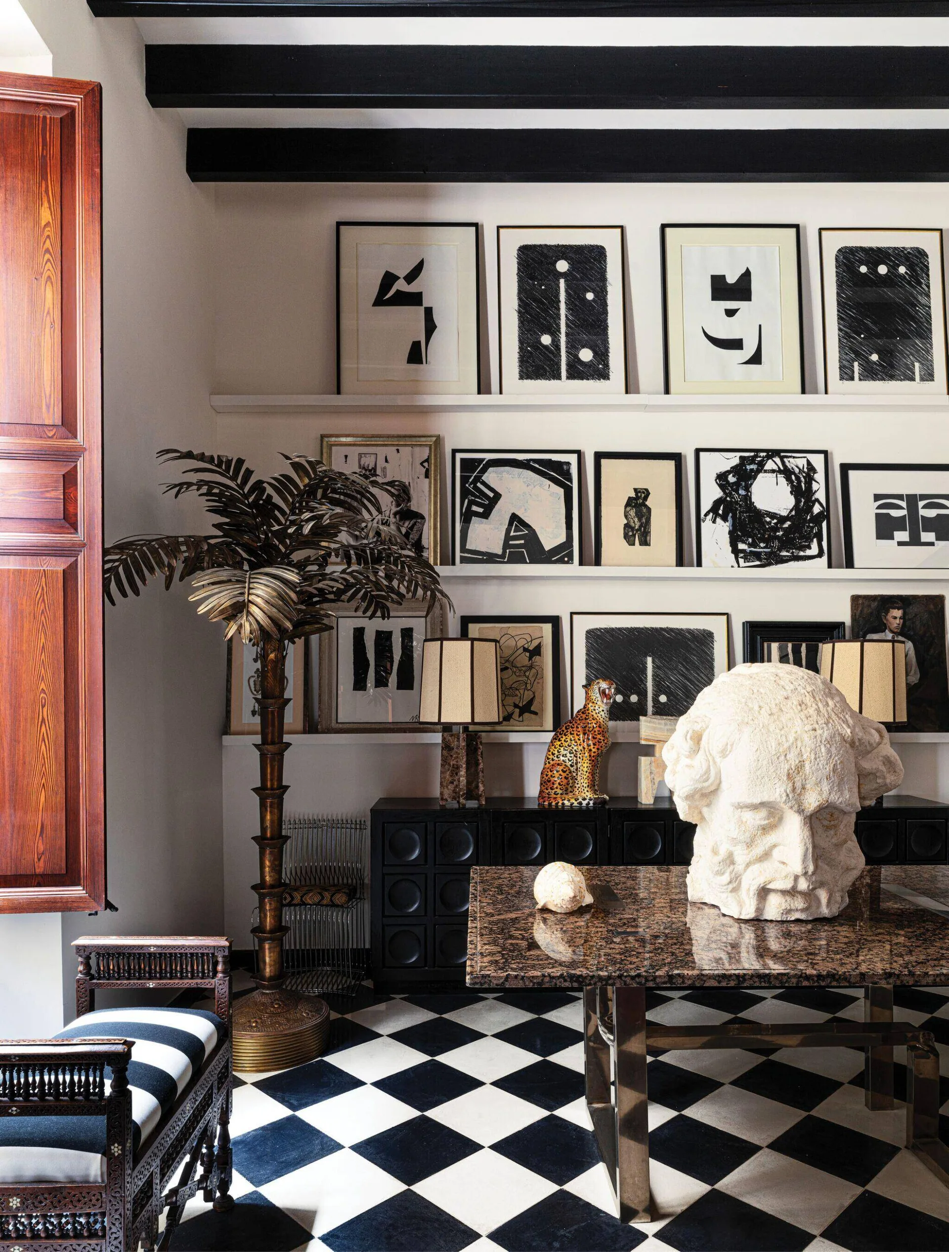 A black and white checkerboard floor anchors an eclectic room featuring a stone sculpture, a brass palm tree lamp, and shelves of framed art.