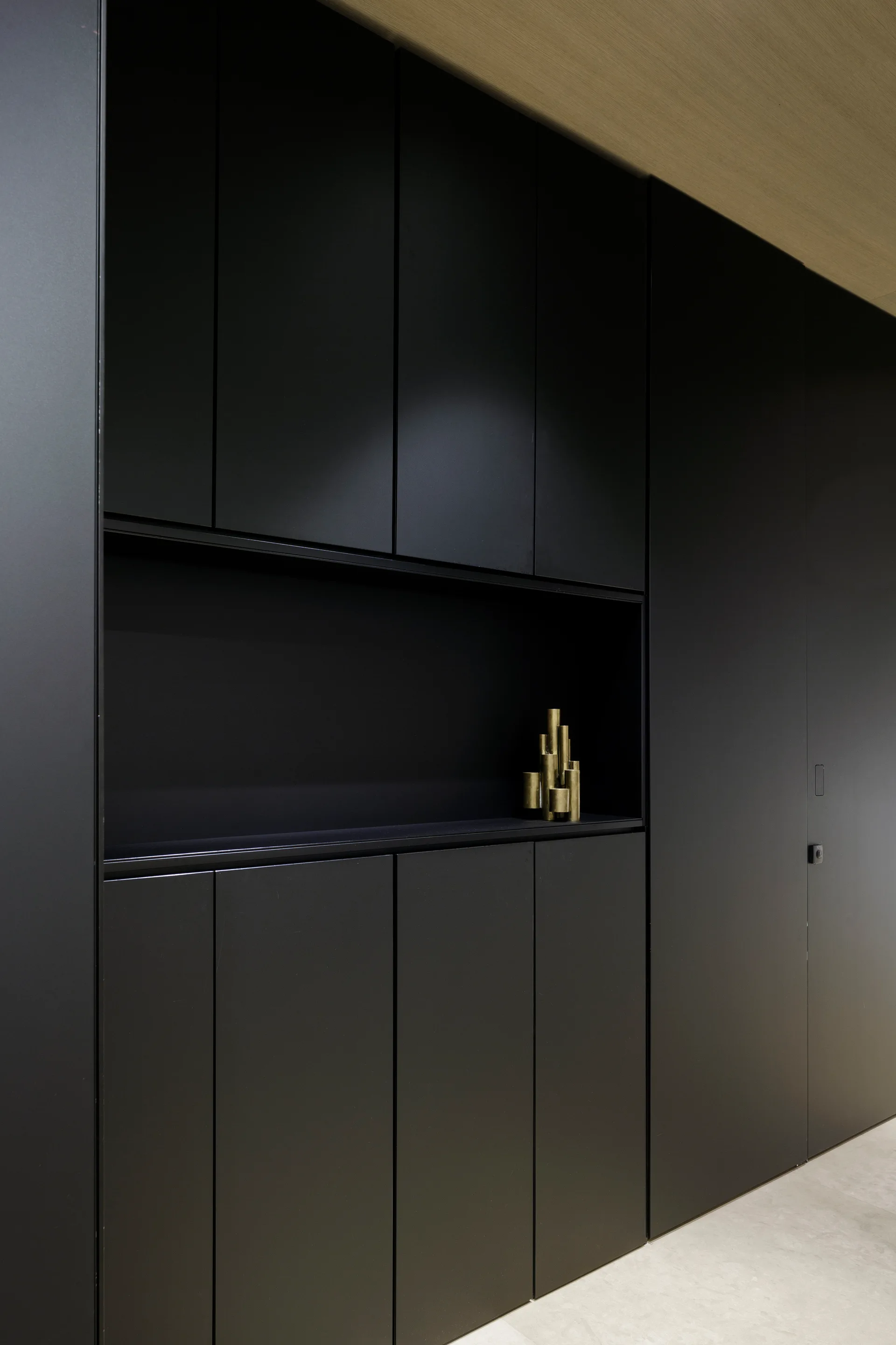 A wall of floor-to-ceiling matte black cabinets with a horizontal recessed open shelf featuring a gold cylindrical sculptural piece.