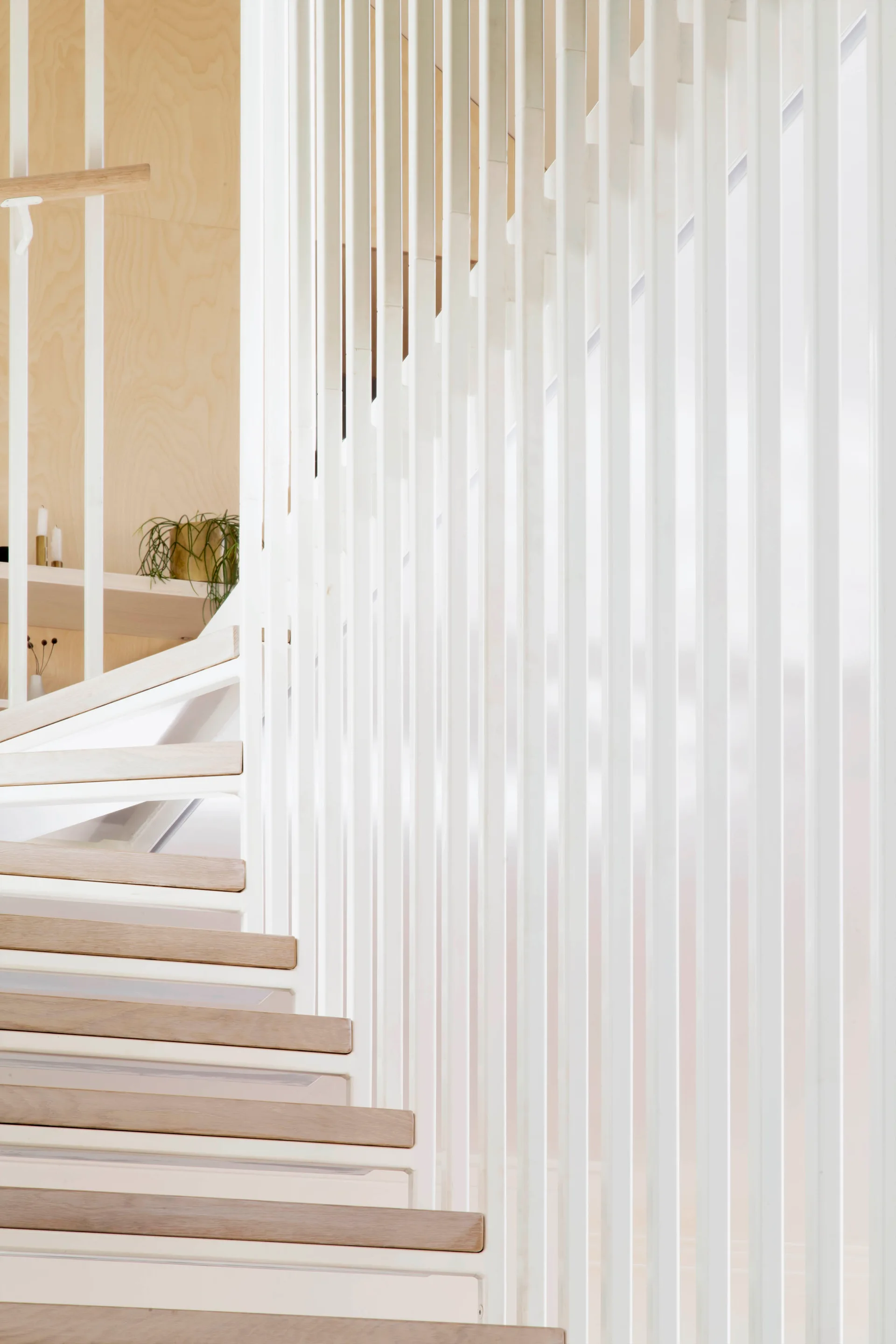 A close-up of a spiral staircase featuring light wood treads and a screen of vertical white metal balusters against a light plywood wall.