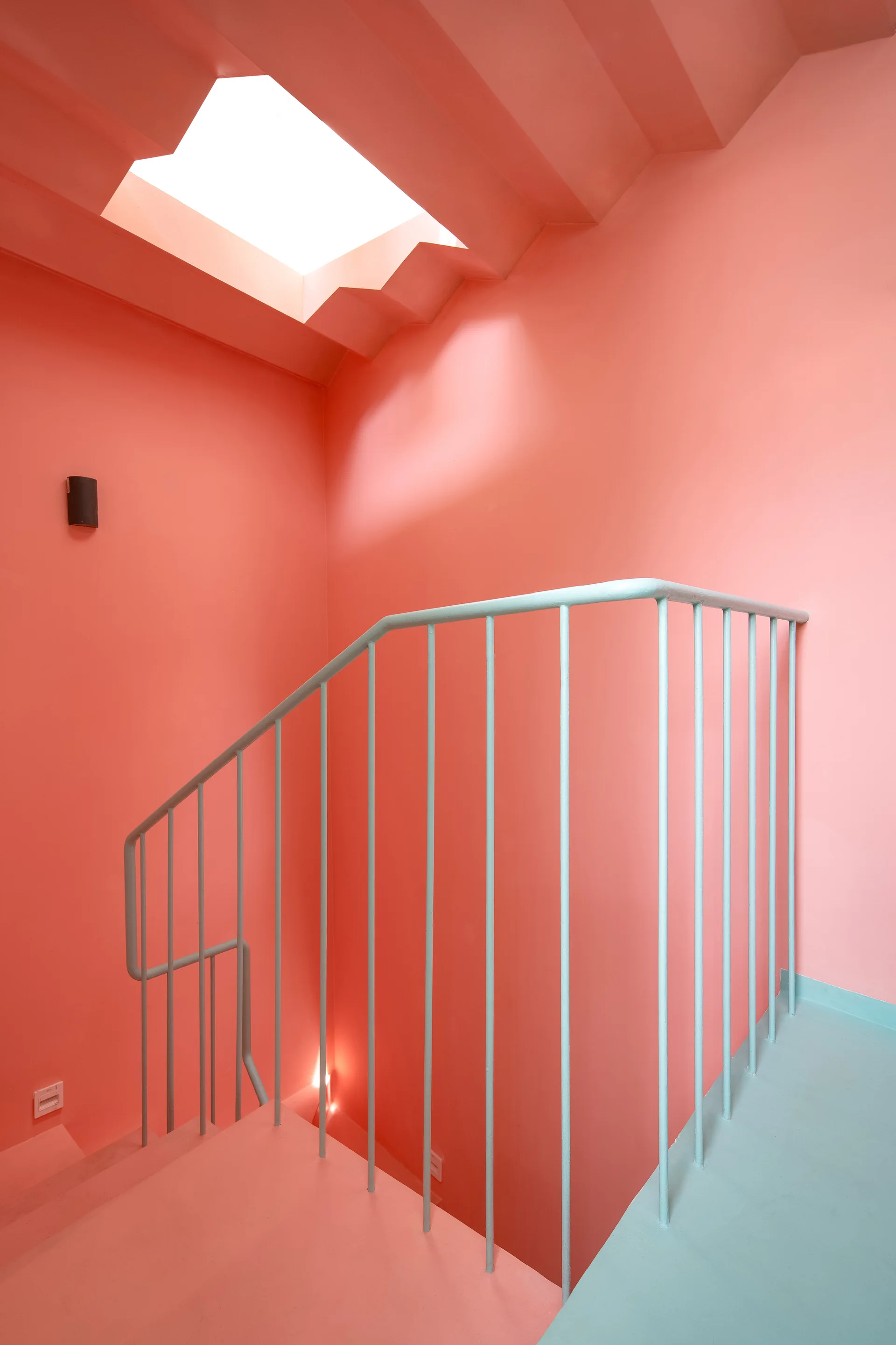 A minimalist staircase features salmon pink painted walls, a light blue metal railing, and a bright rectangular skylight overhead.