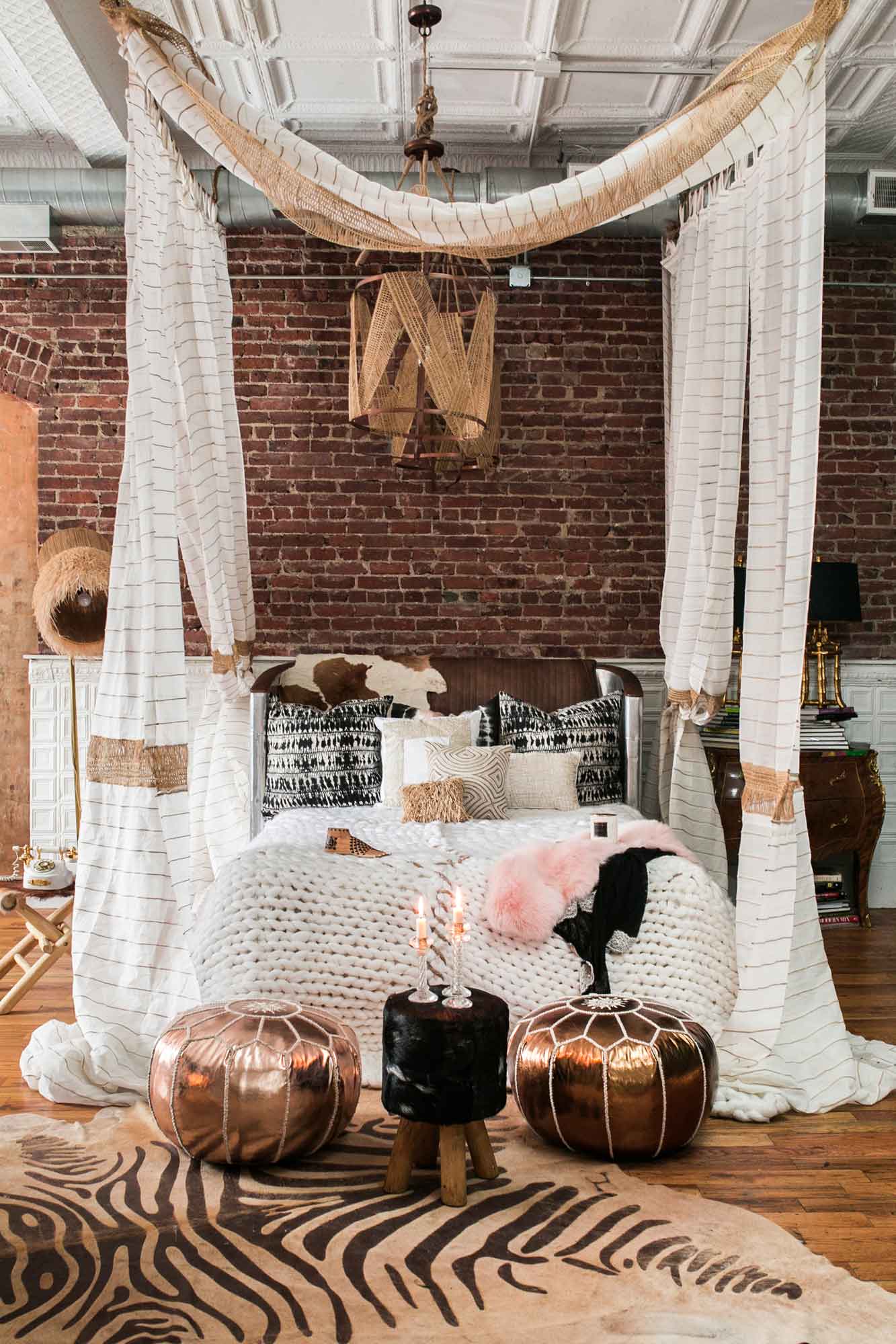 A draped canopy bed with patterned pillows sits against an exposed brick wall, with metallic poufs and a zebra print rug on the wood floor.