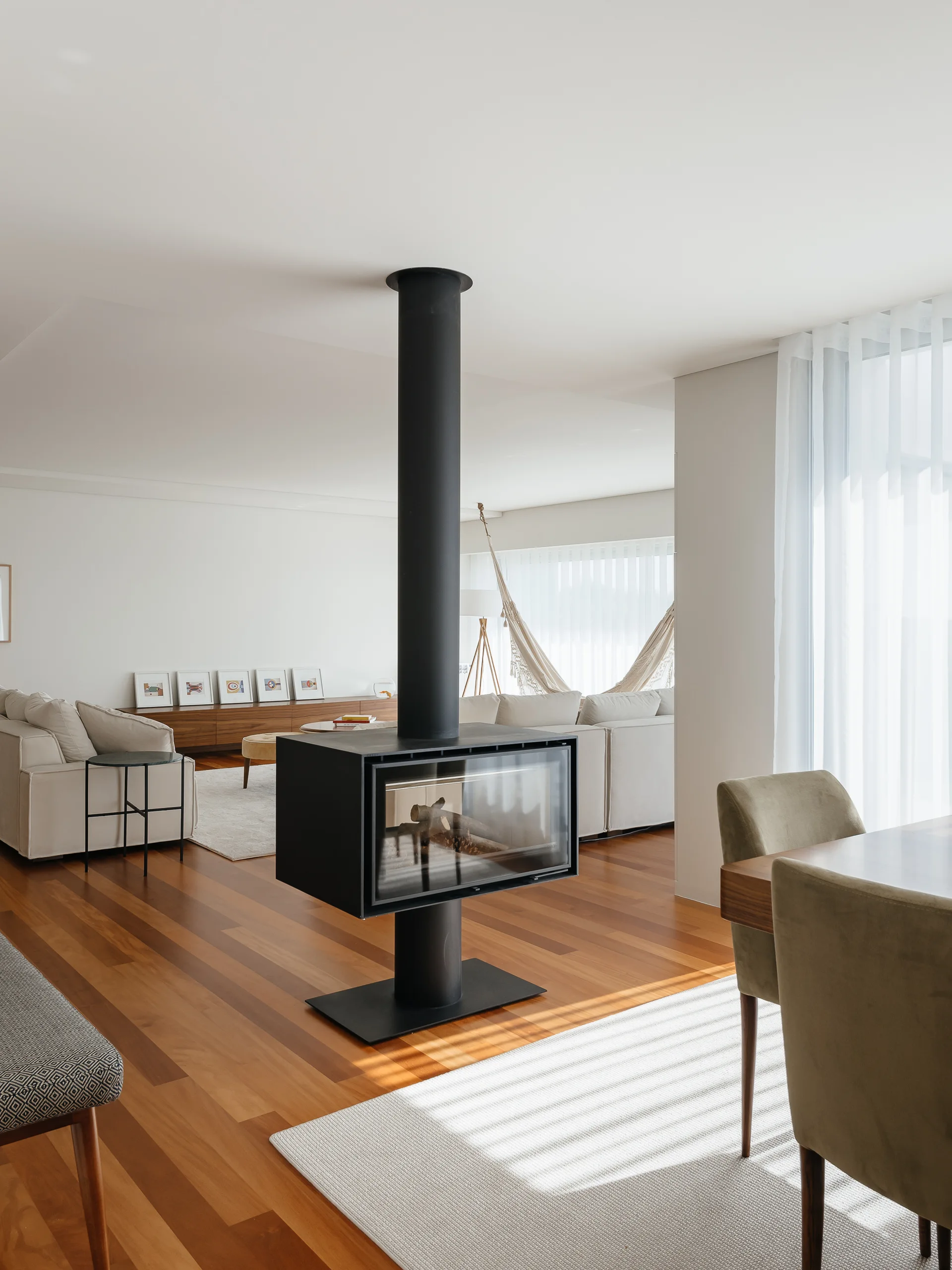 An open living and dining space featuring a central black freestanding fireplace, hardwood floors, a modular beige sofa, and sheer curtains.