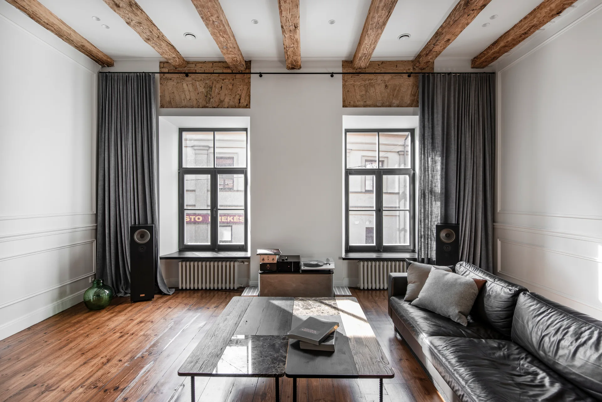 A bright living room features exposed wood ceiling beams, a black leather sofa, two large windows with gray curtains, and polished wood floors.