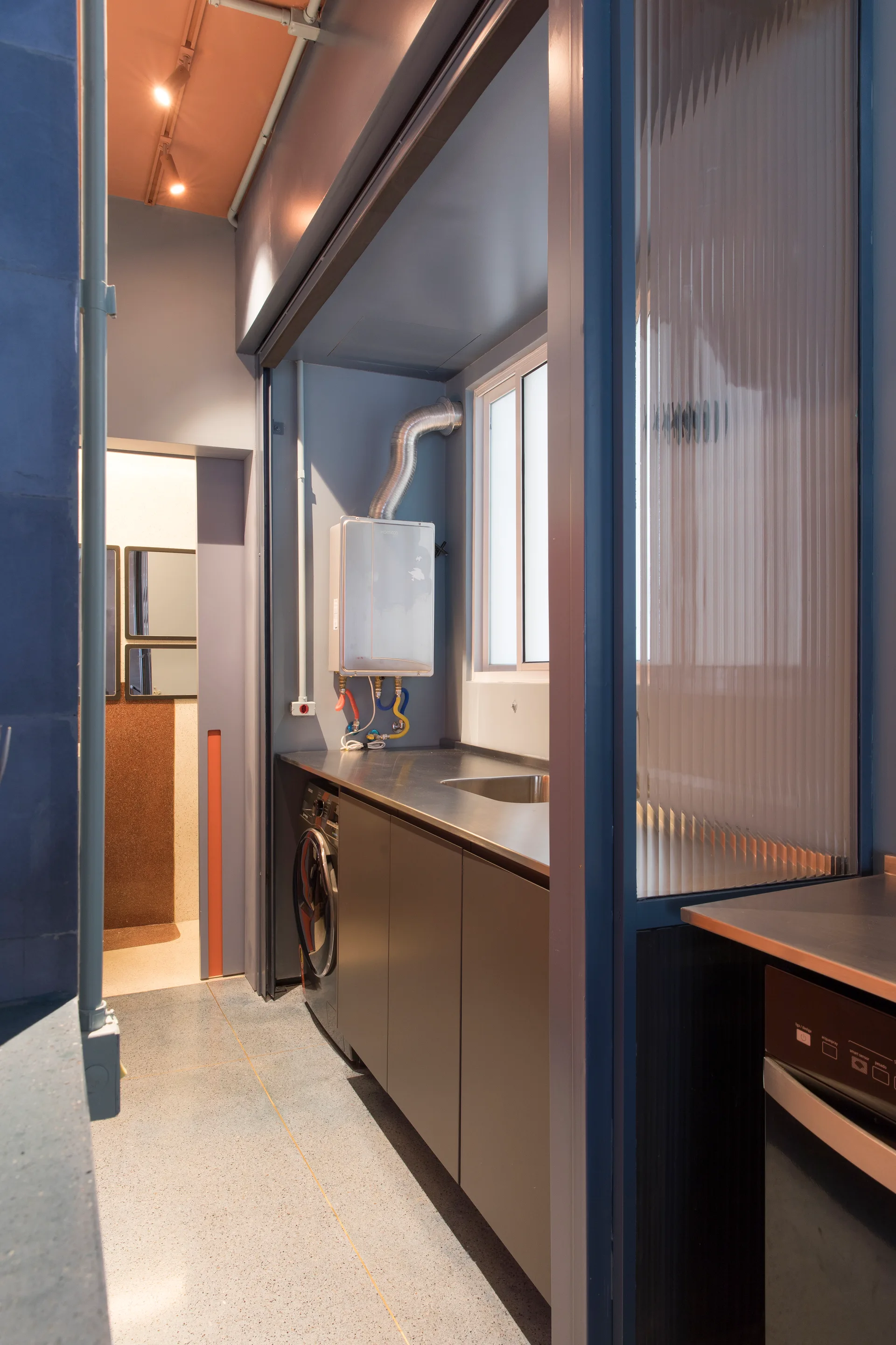A modern utility laundry room with a stainless steel countertop, a washing machine, and a blue-framed fluted glass partition screen.