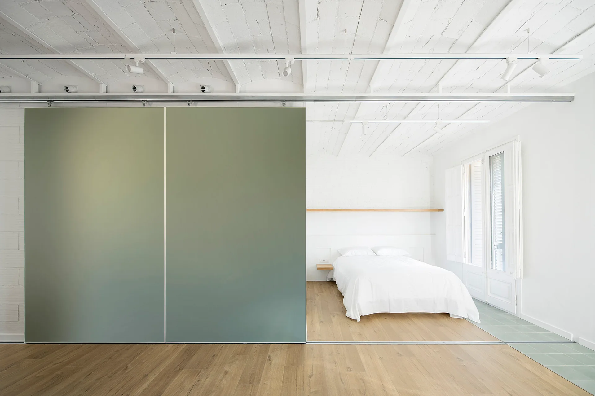 A minimalist white bedroom featuring a white bed, exposed white ceiling beams, and a large sage green sliding partition door.