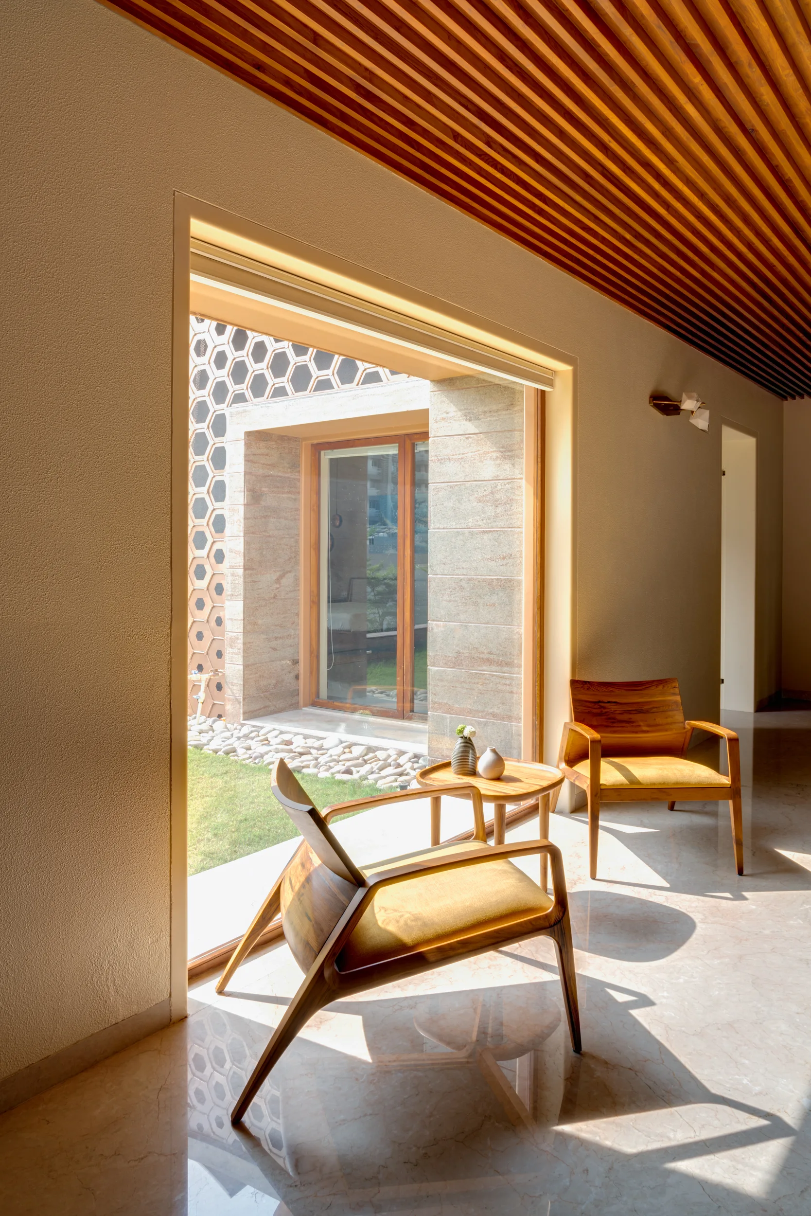 Two wooden armchairs with yellow cushions sit on a polished marble floor beneath a slatted wood ceiling next to a large courtyard window.