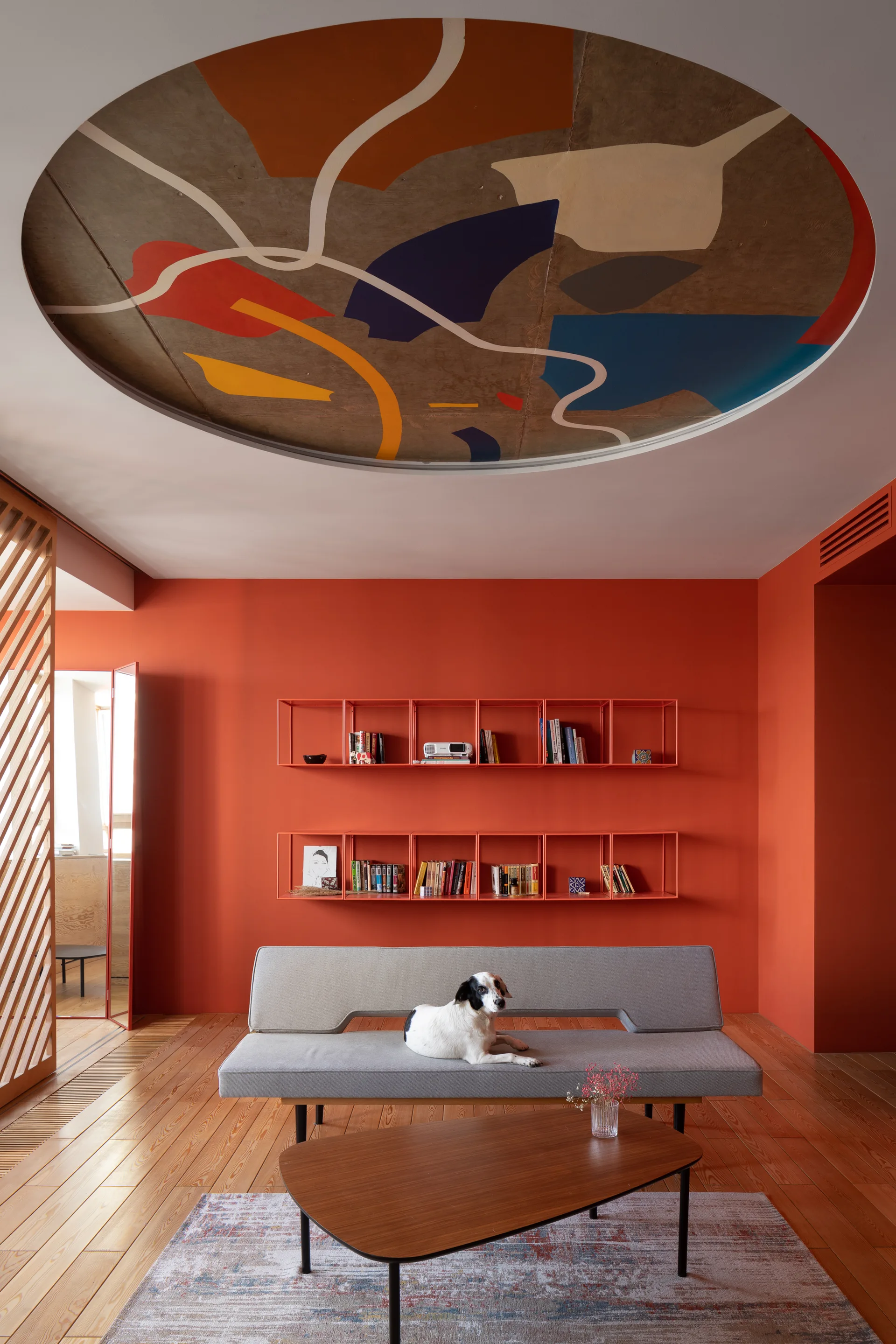 A minimalist grey sofa sits against a bright orange wall in a living room featuring an abstract circular ceiling mural and wooden floors.
