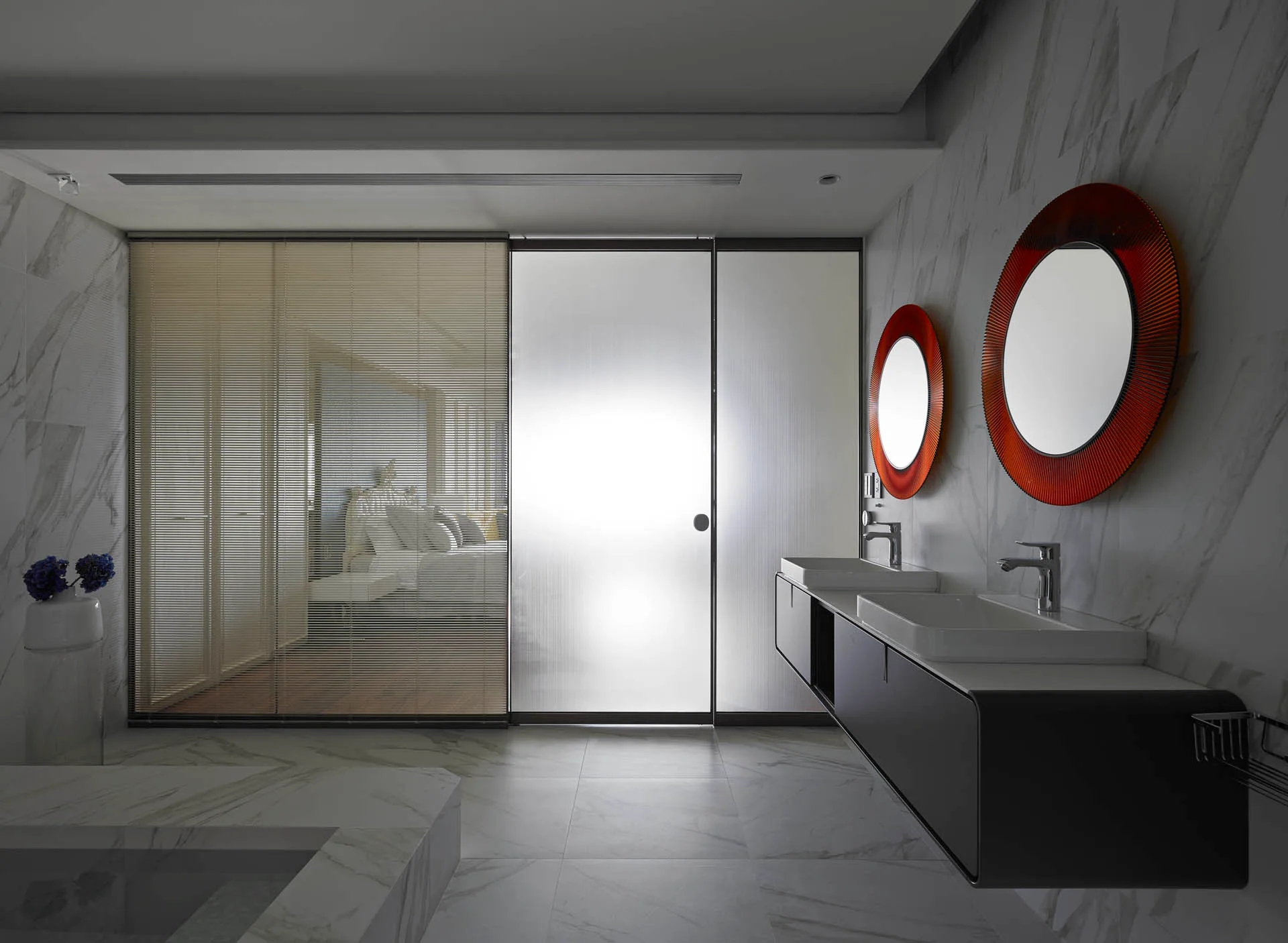 A modern marble bathroom features a dark floating vanity with dual sinks below two round orange-framed mirrors next to a sliding glass partition.