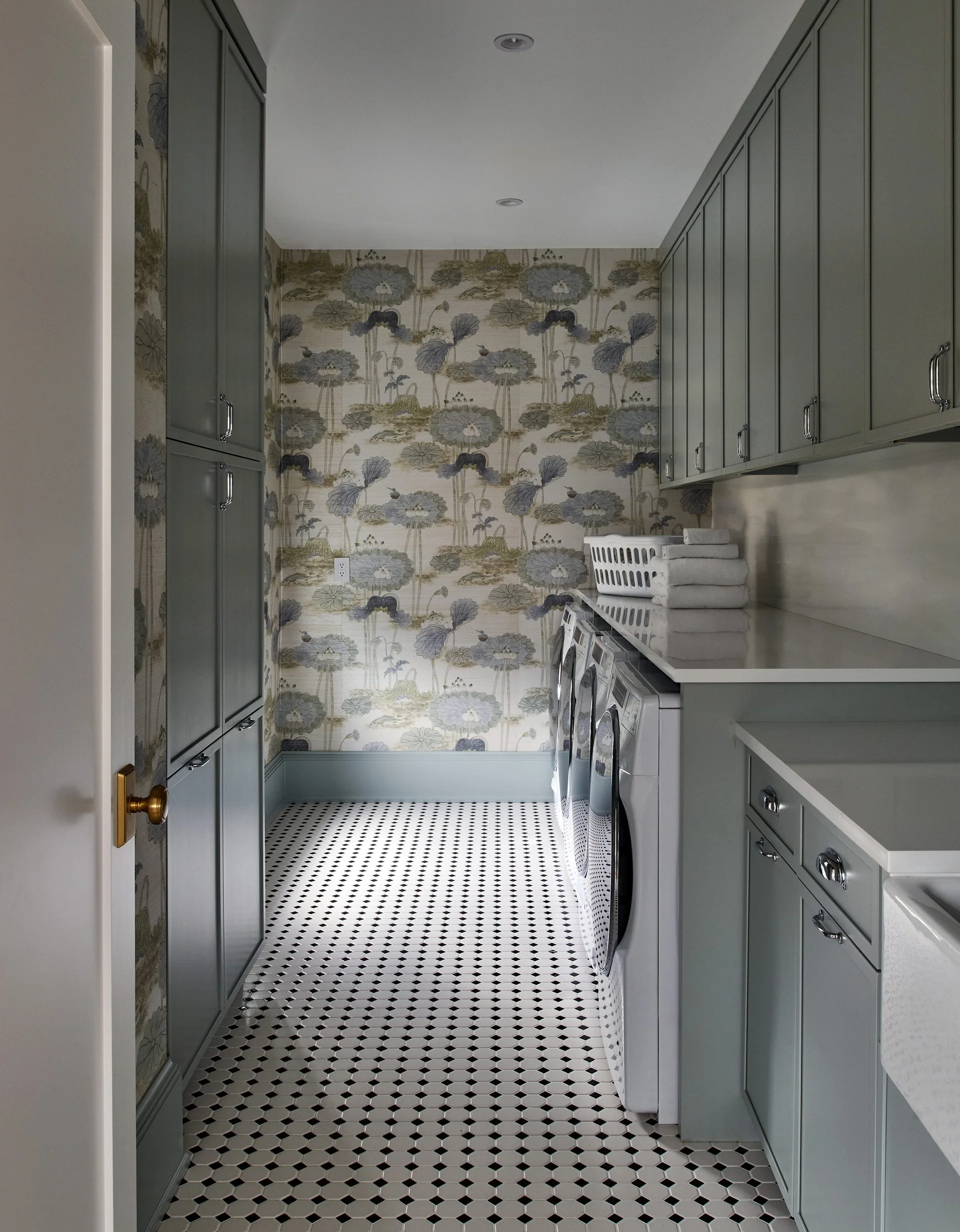 A narrow laundry room features sage green cabinets, botanical wallpaper on the back wall, and a black-and-white geometric tiled floor.
