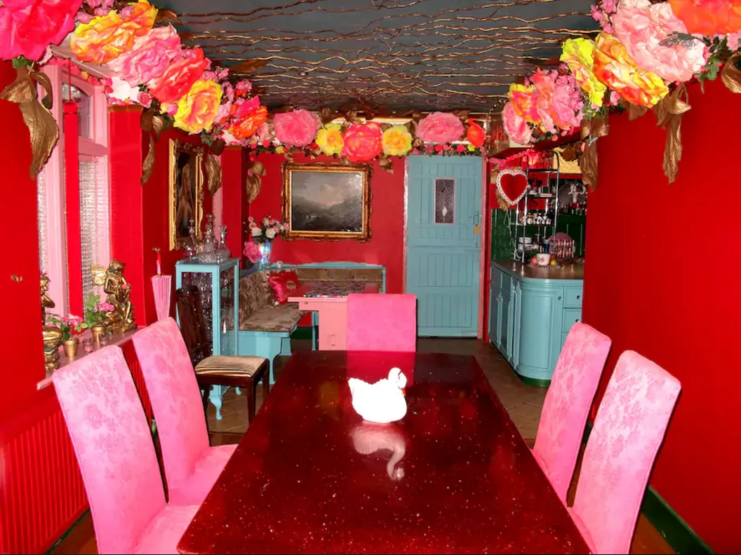 An eclectic dining room features a glossy red dining table, bright pink upholstered chairs, vibrant red walls, and a light blue painted door.