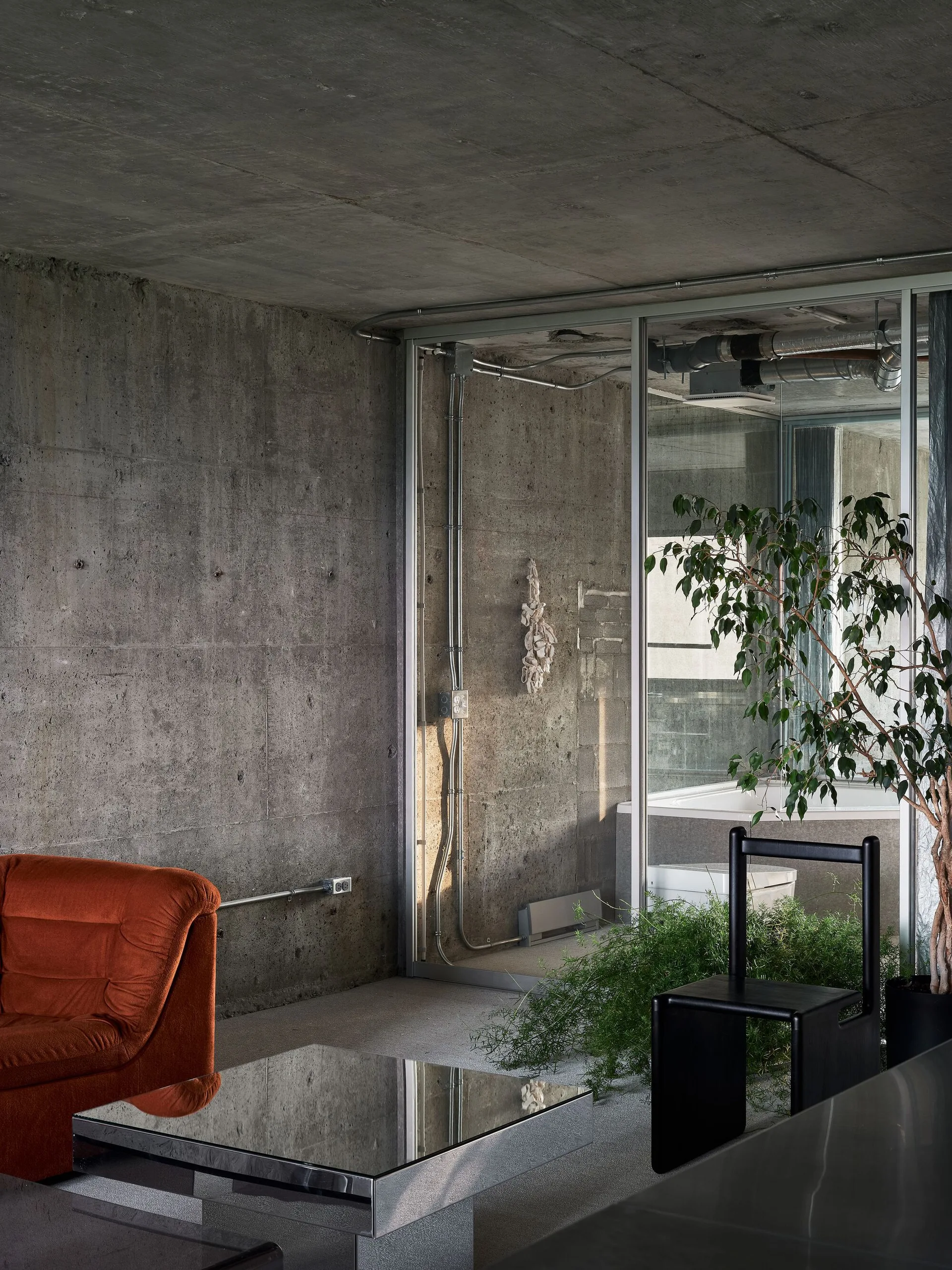 An industrial living room features raw concrete walls, a terracotta-orange sofa, a mirrored coffee table, and a glass partition enclosing a bathroom.
