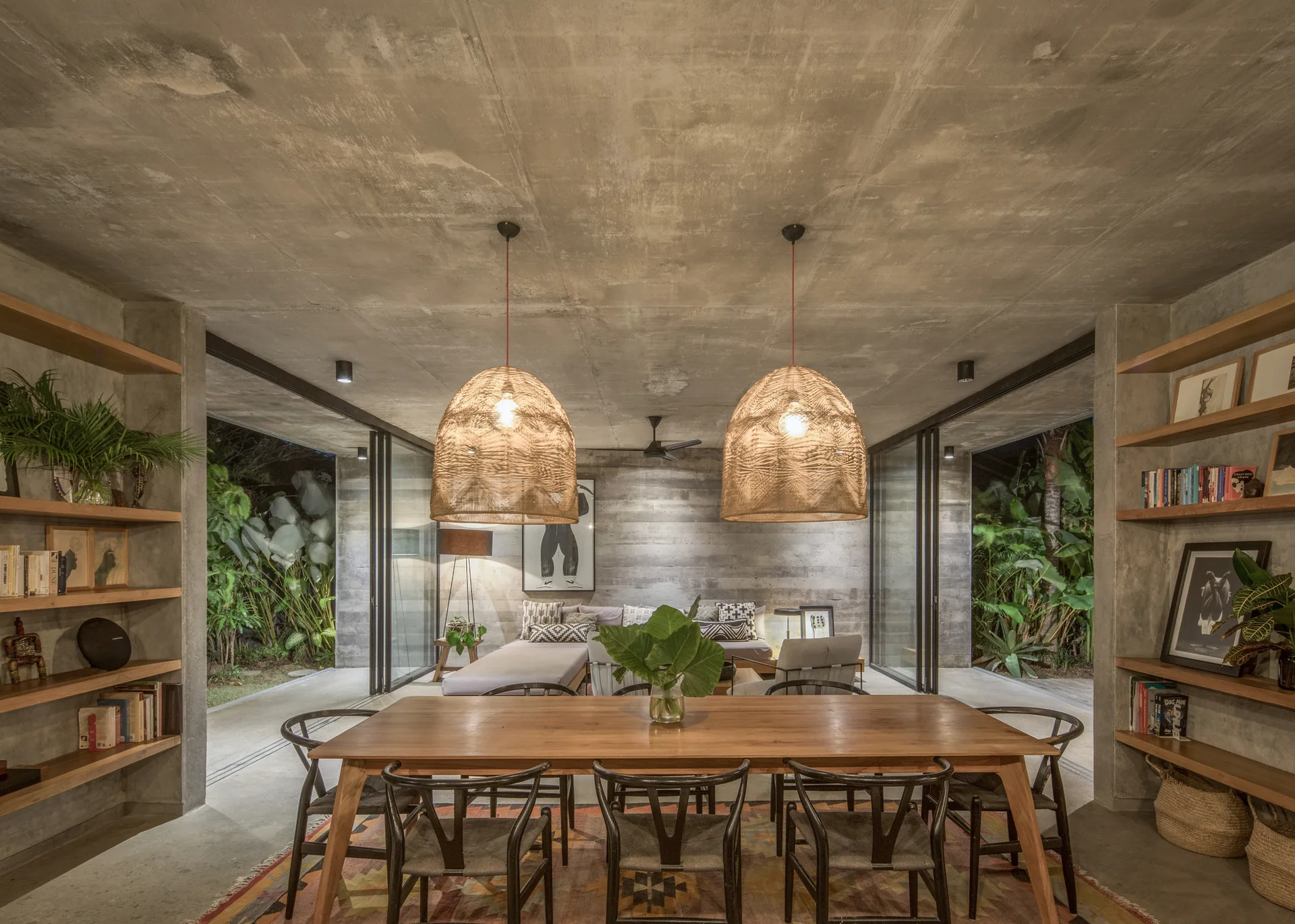 A dining room with raw concrete walls features a wooden table, black wishbone chairs, and two large woven pendant lights hanging from the ceiling.