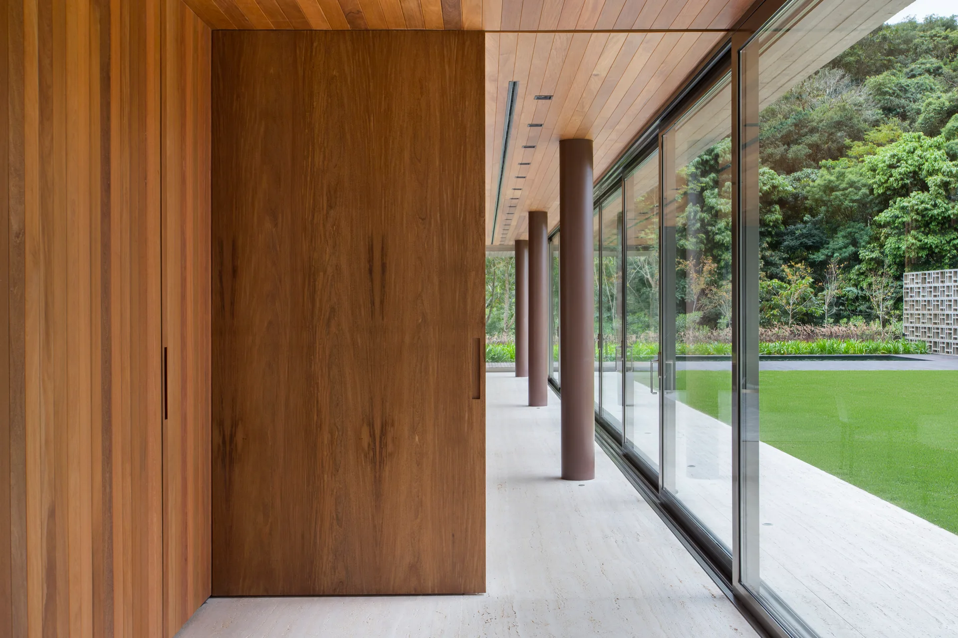 A minimalist hallway features warm wood wall and ceiling paneling, light stone flooring, and large glass sliding doors looking out to a green lawn.