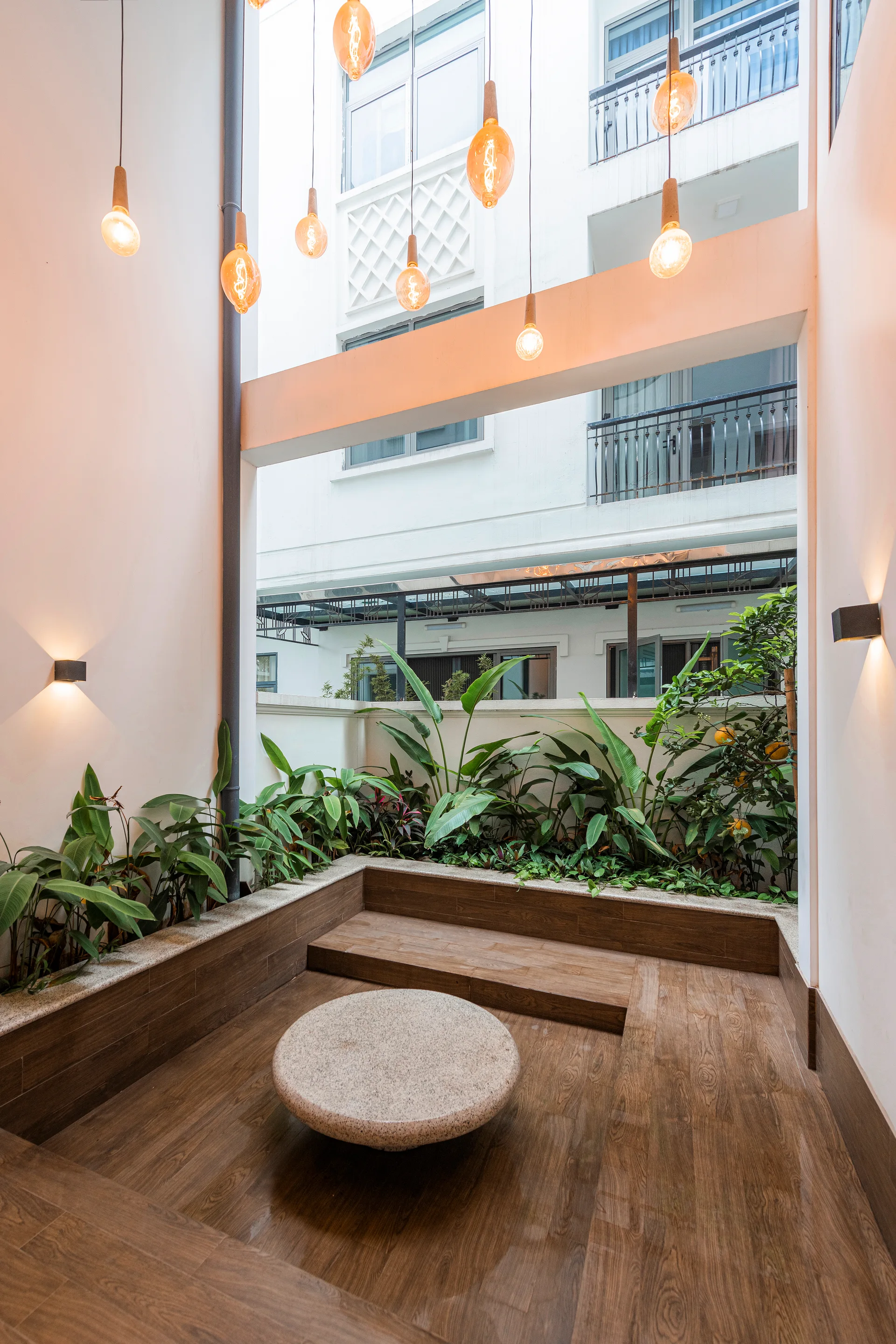 An open-air courtyard features tiered wooden decking, built-in planters with green plants, suspended filament lightbulbs, and a round stone table.