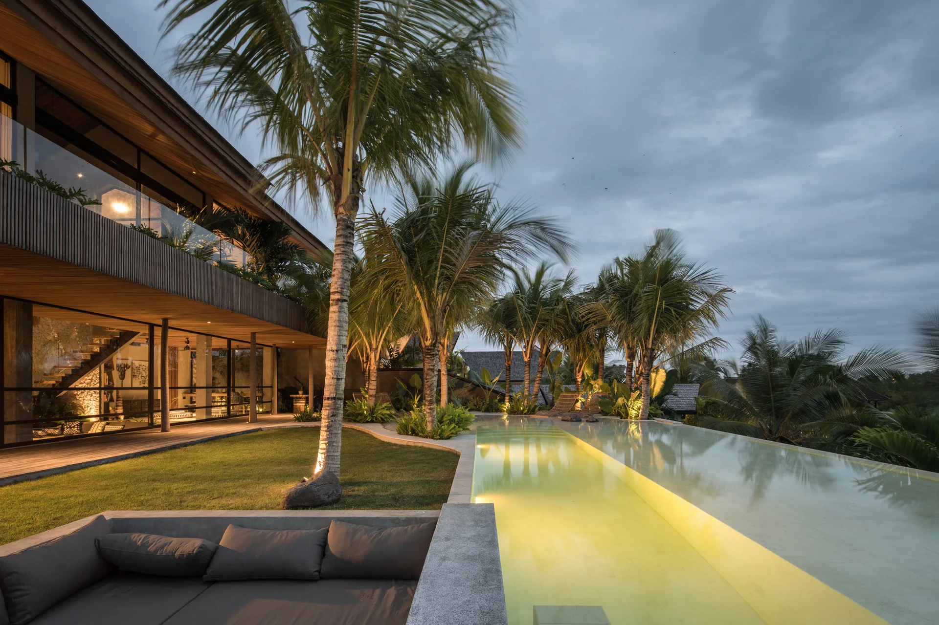 An infinity pool glowing with warm light sits next to a modern wood-clad house, flanked by palm trees and low concrete lounge seating.