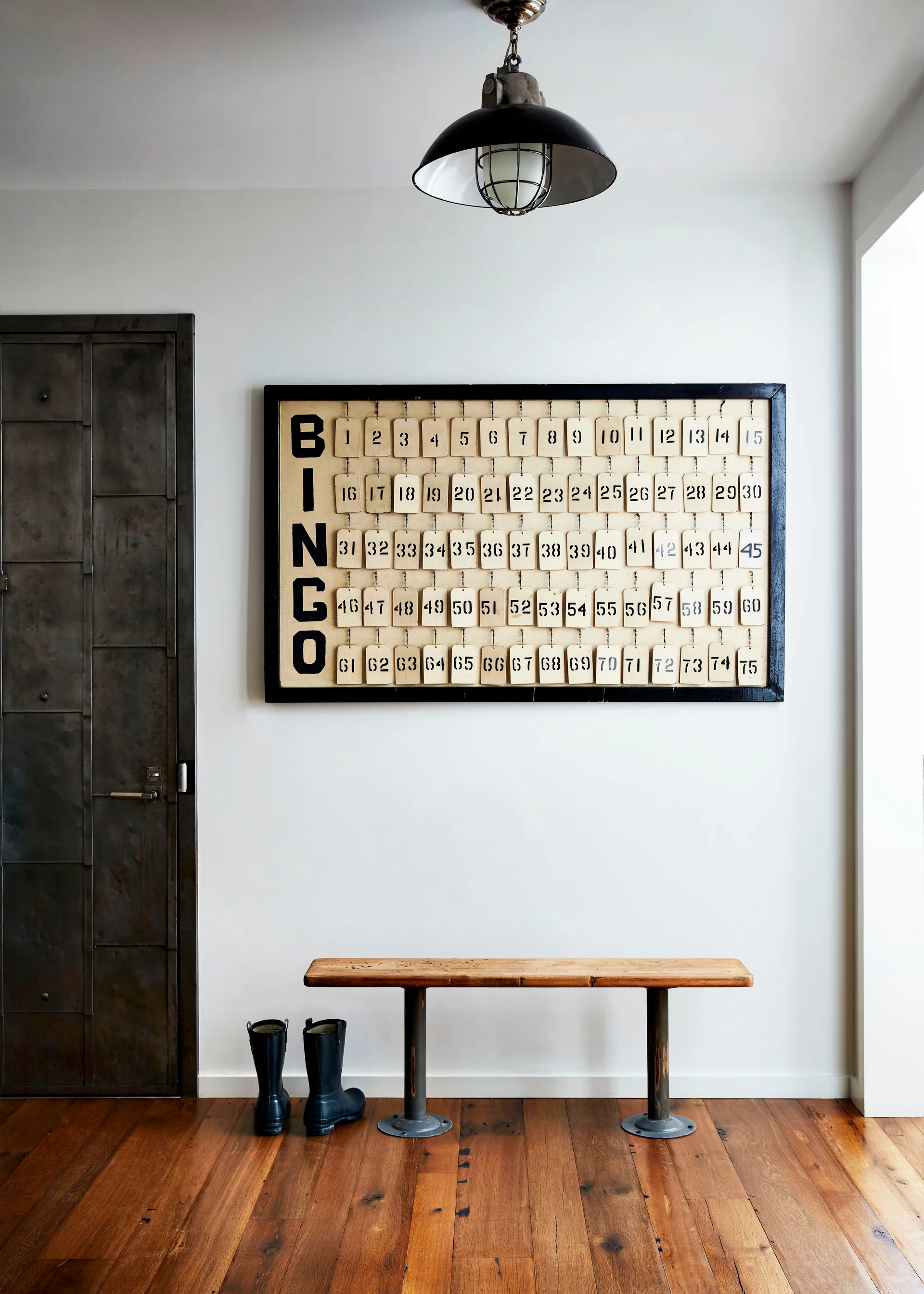 An industrial entryway features a wooden bench, a pair of black rain boots, a metal door, and a large vintage bingo board on a white wall.