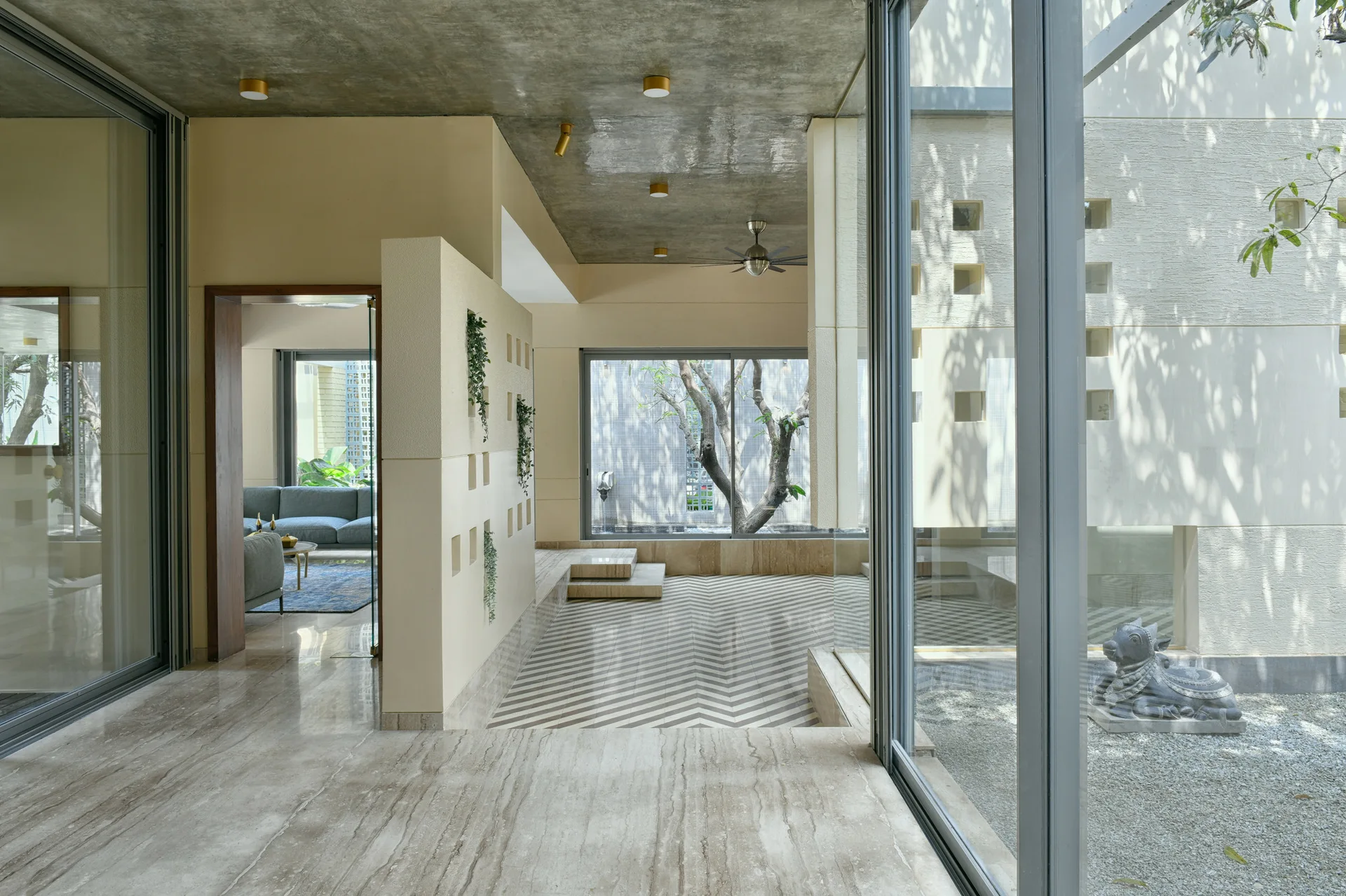 A modern interior corridor features a textured concrete ceiling, a cream partition wall with square cutouts, and a chevron-patterned stone floor.