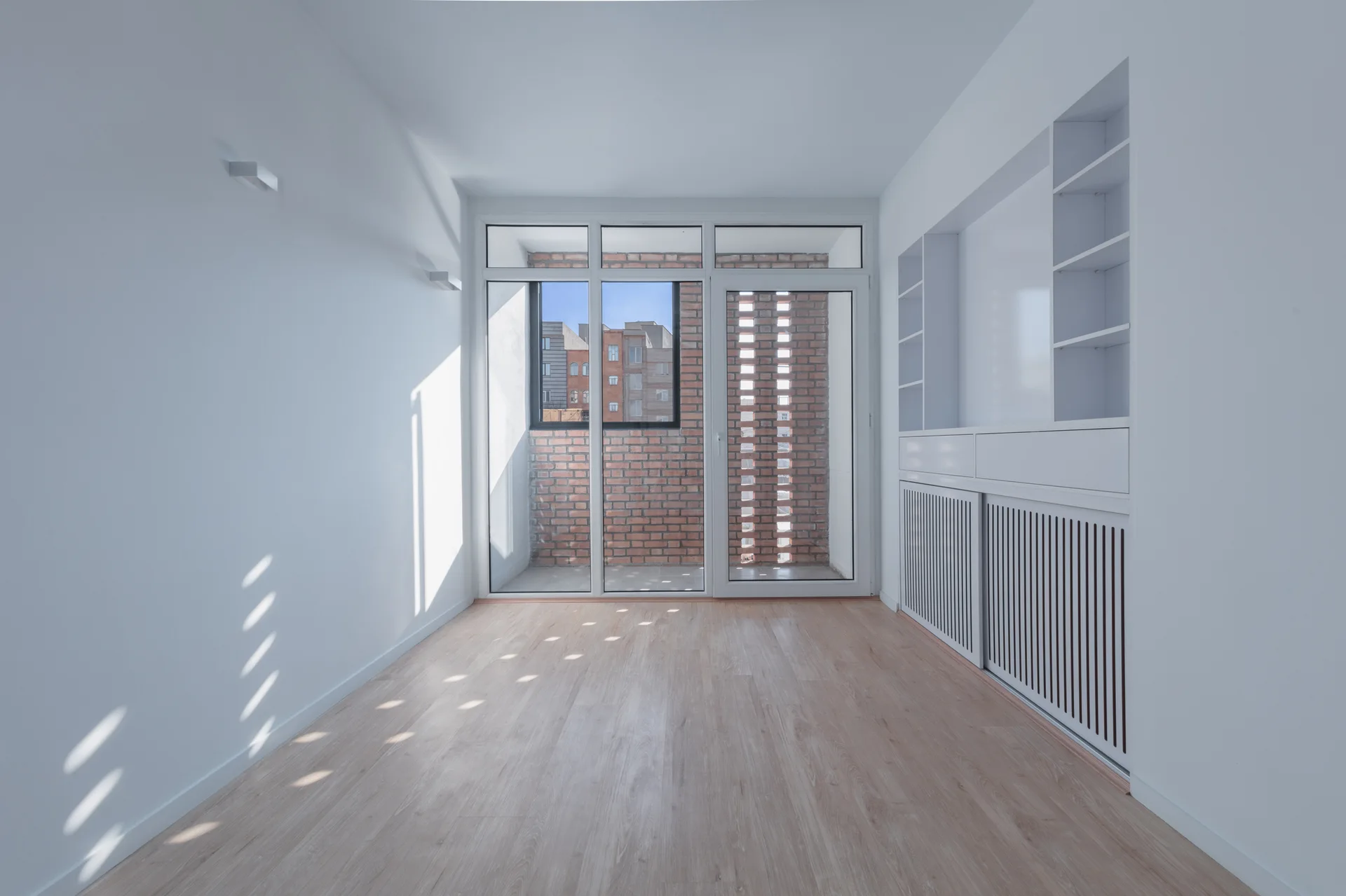 An empty minimalist room features light wood flooring, white walls, custom built-in cabinetry, and a glass door leading to a brick balcony.