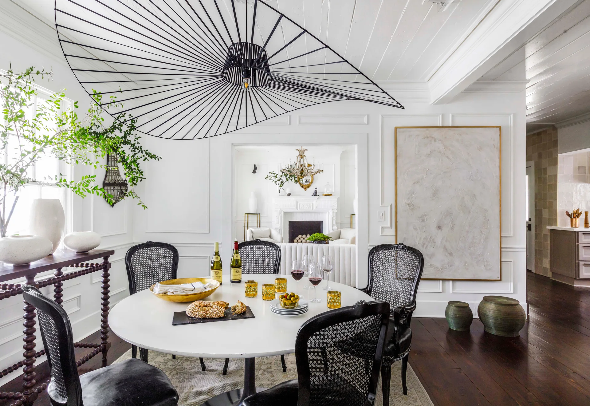 A bright white dining room features a round table, black cane chairs, and a large black wire pendant light under a wood plank ceiling.