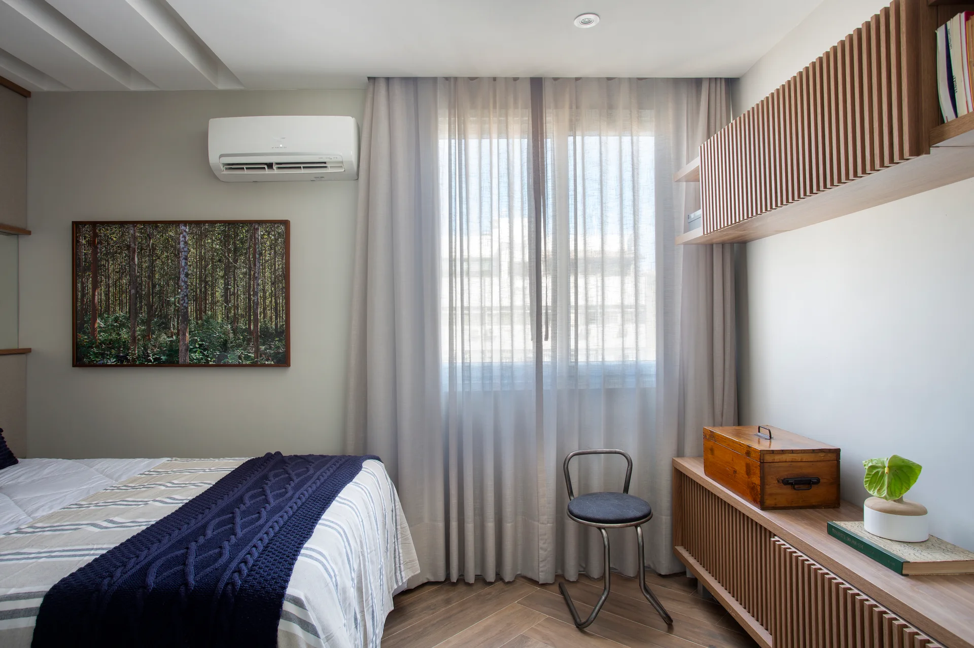 A contemporary bedroom featuring a bed with striped linen sheets, slatted wood storage cabinets, and sheer grey curtains.