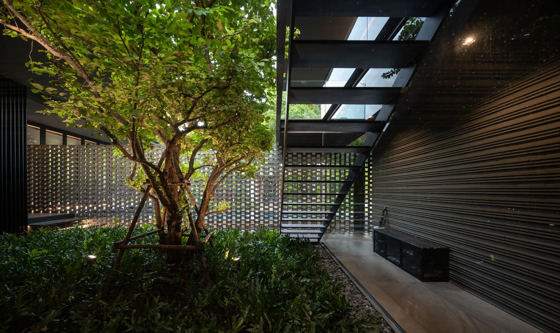 An architectural outdoor courtyard with a central tree, an open-riser black metal staircase, a horizontal slatted wall, and a breeze-block screen.