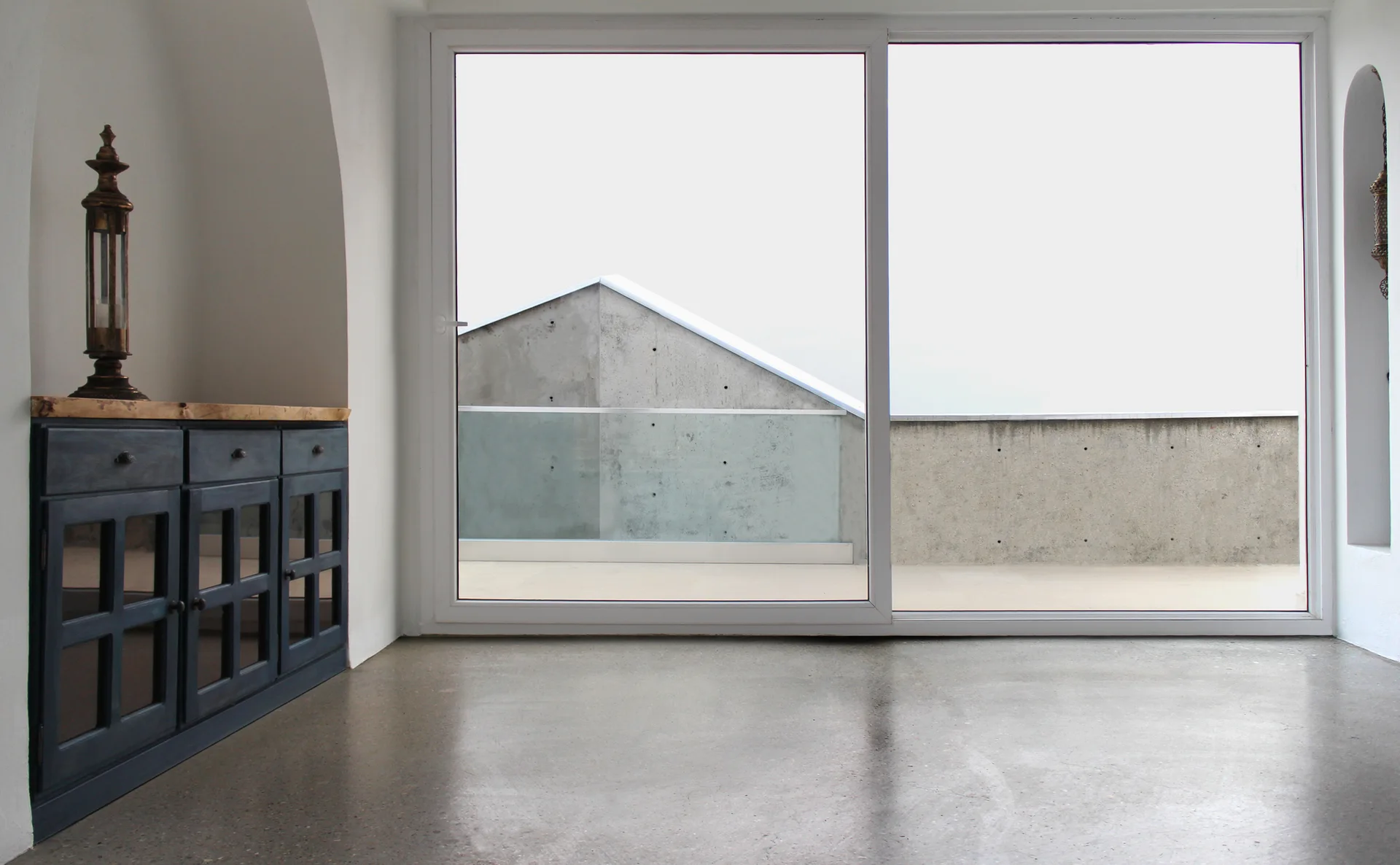 A minimalist room features a dark blue wooden cabinet, polished grey concrete flooring, and a large sliding glass door opening to a concrete balcony.
