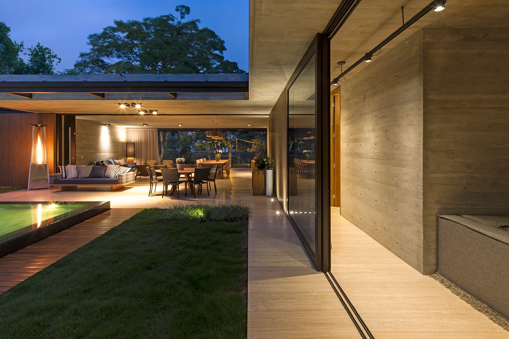 An open-concept outdoor living area at twilight features a swimming pool, a wood deck, and a lounge space framed by a board-formed concrete wall.