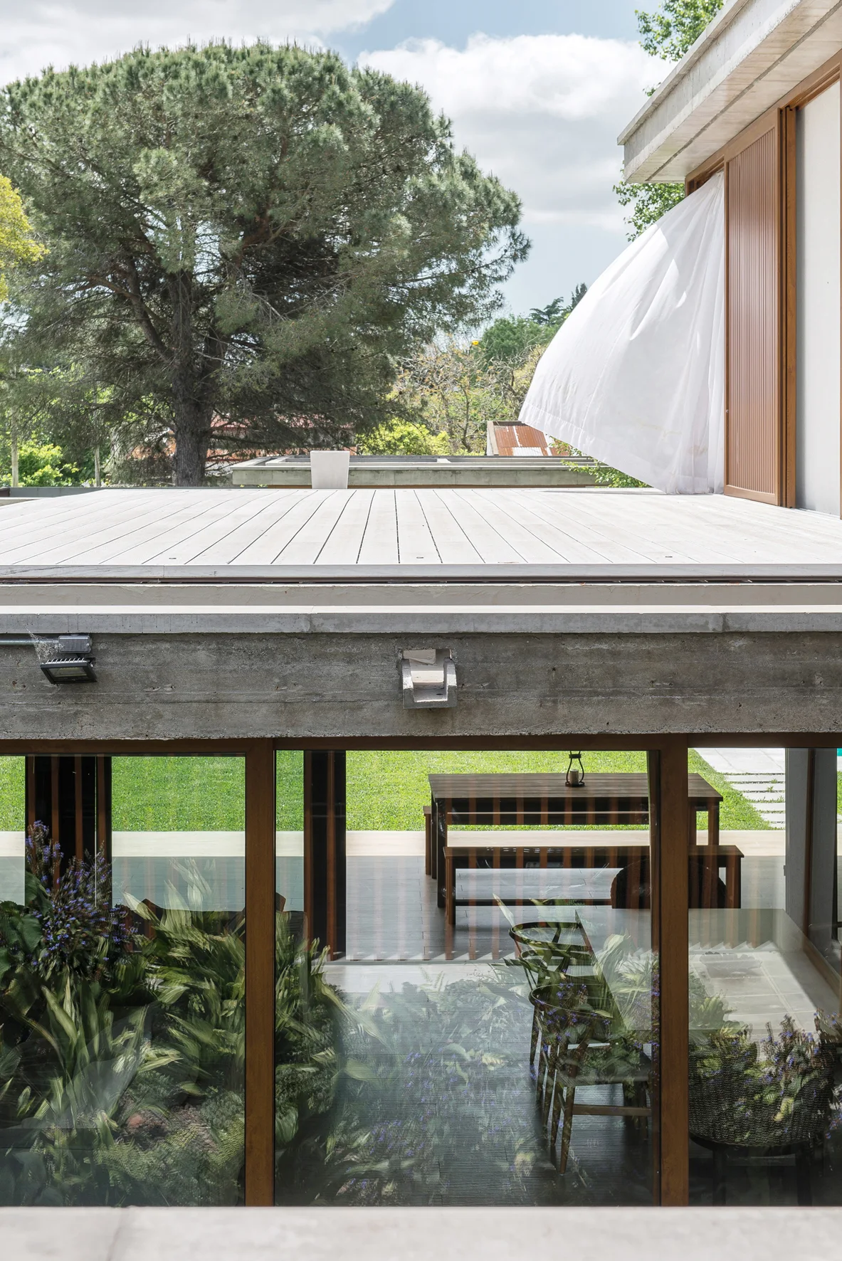 An elevated wooden deck sits above a concrete-framed patio with glass doors showing an indoor garden and outdoor dining table.