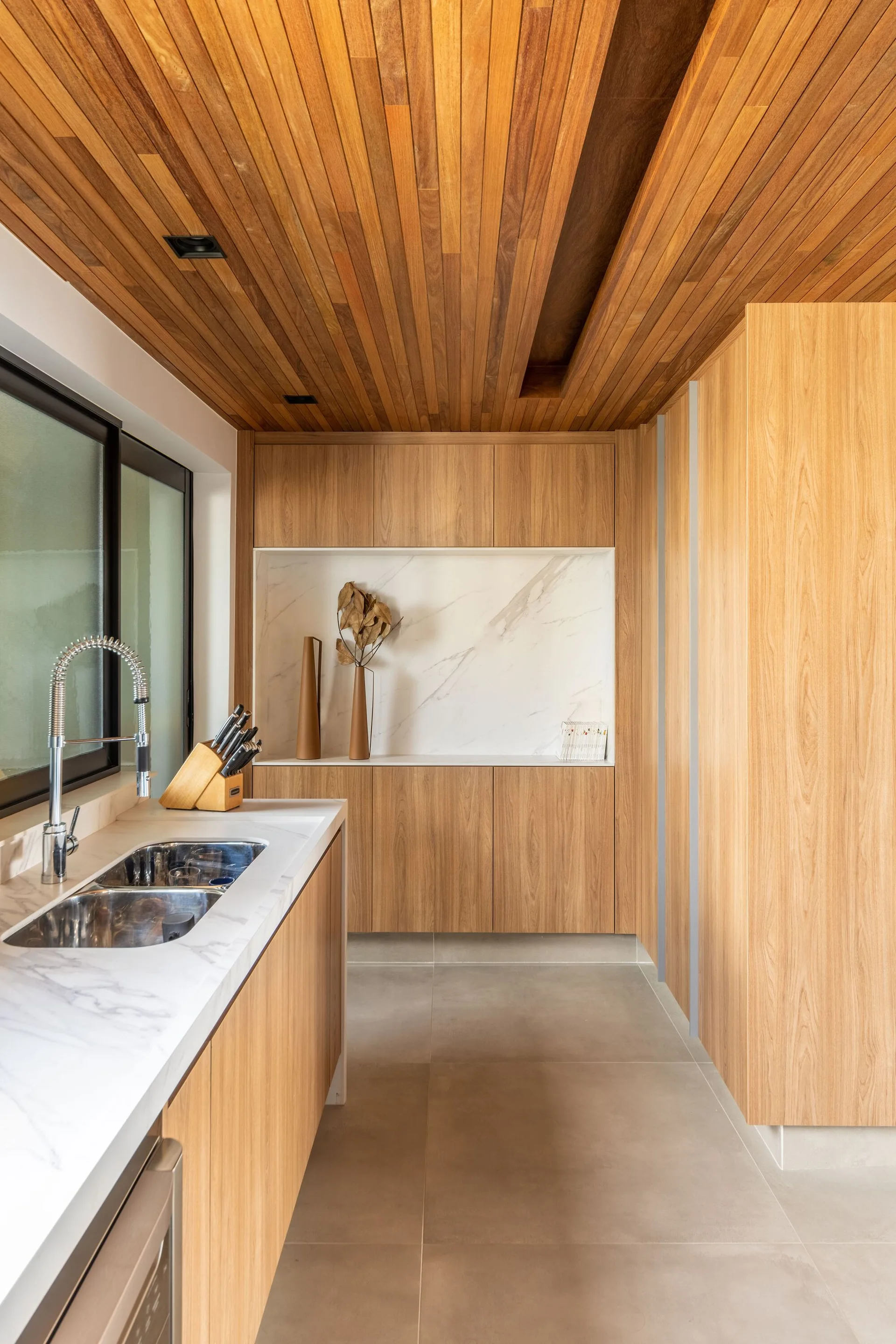 A modern kitchen features light wood cabinetry, a matching slatted wood ceiling, grey floor tiles, and a white marble countertop with a double sink.