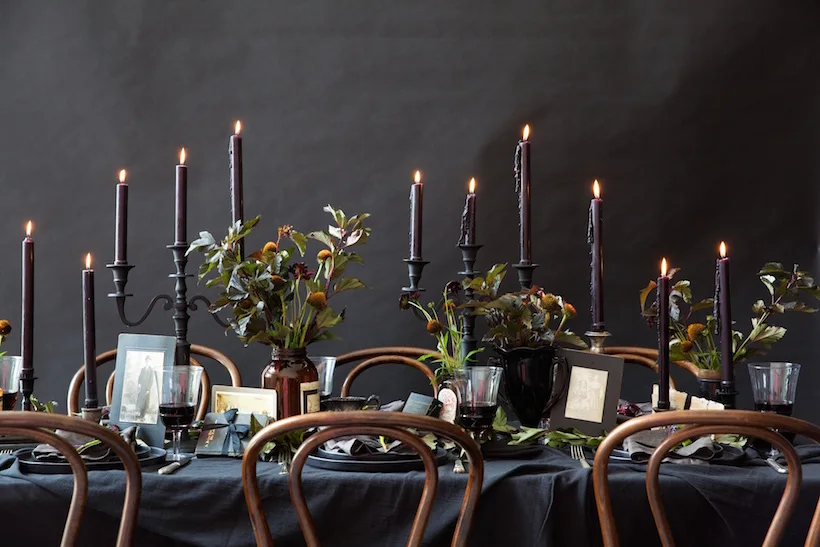 A dark dining table set with a black tablecloth, burning black taper candles, vintage framed photographs, and bentwood chairs against a black wall.