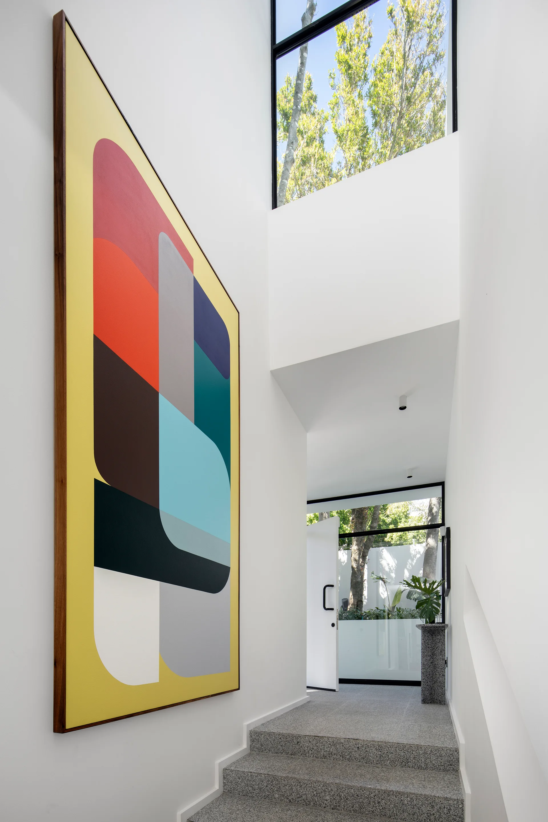 A white minimalist staircase features speckled stone steps, a large colorful geometric painting on the wall, and a high window looking out at trees.