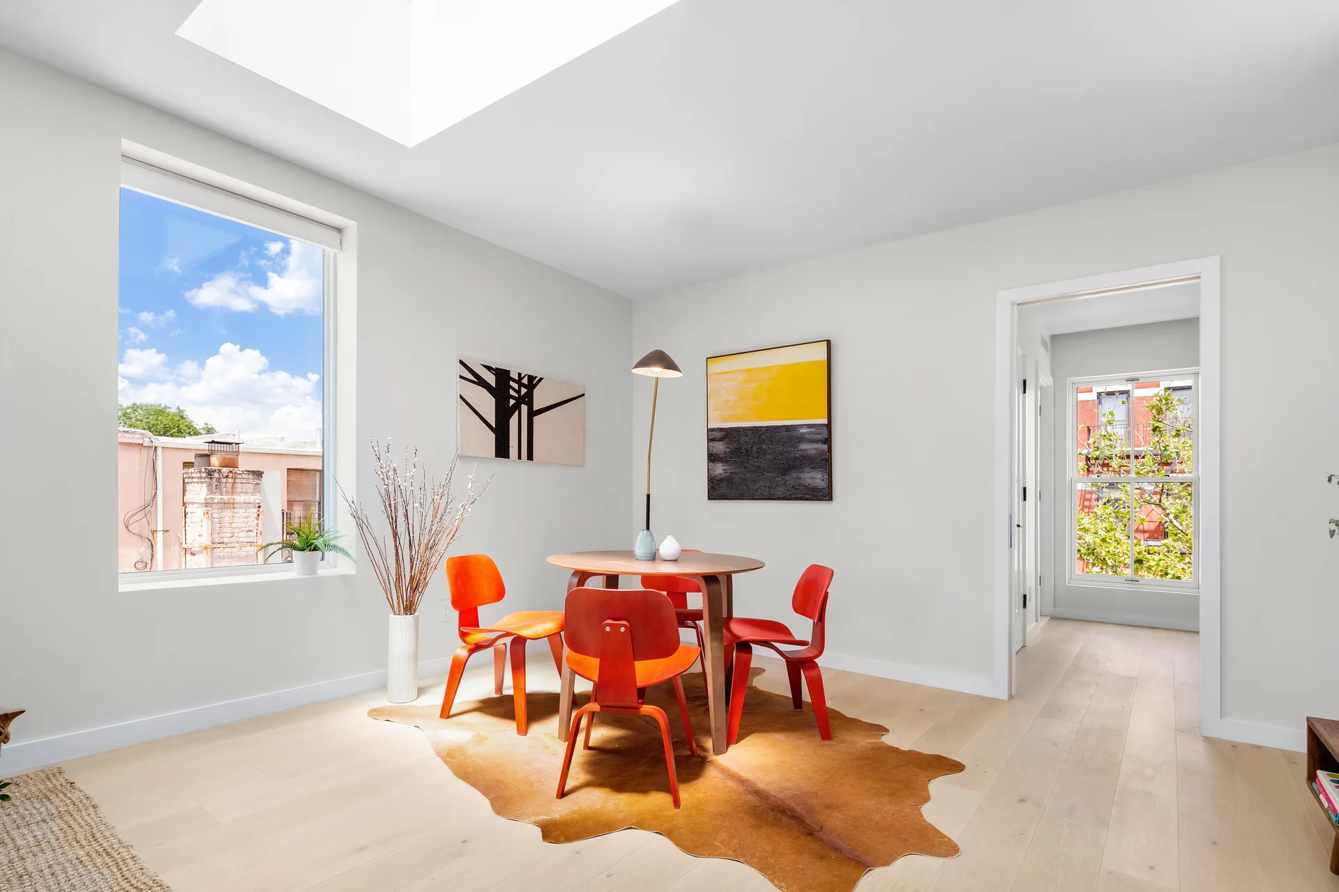A minimalist dining area features a round wood table, orange bentwood chairs, a brown cowhide rug, and natural light from a skylight.