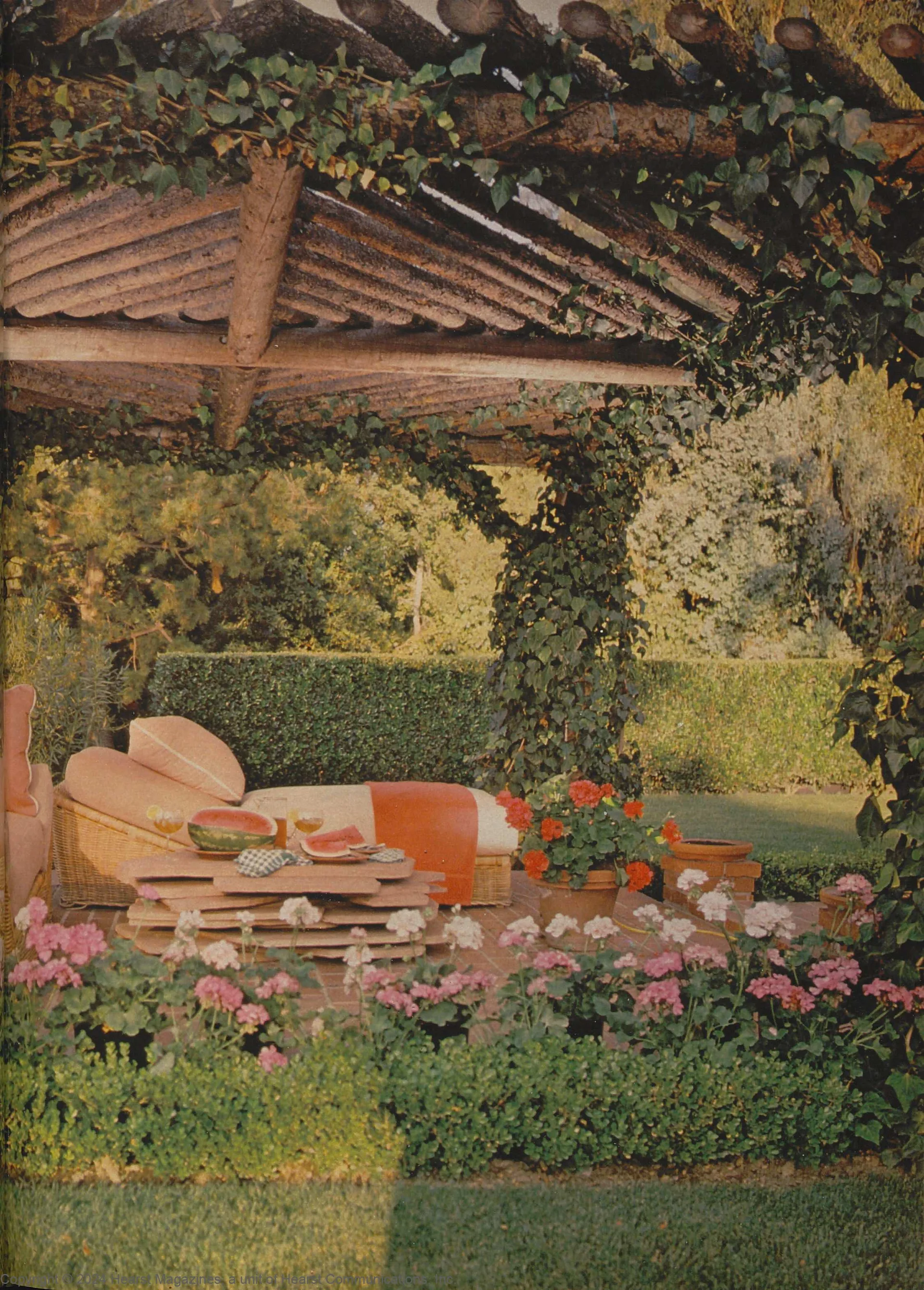 A wicker daybed sits under a rustic log pergola covered in climbing green ivy next to a stacked stone table on a brick patio.