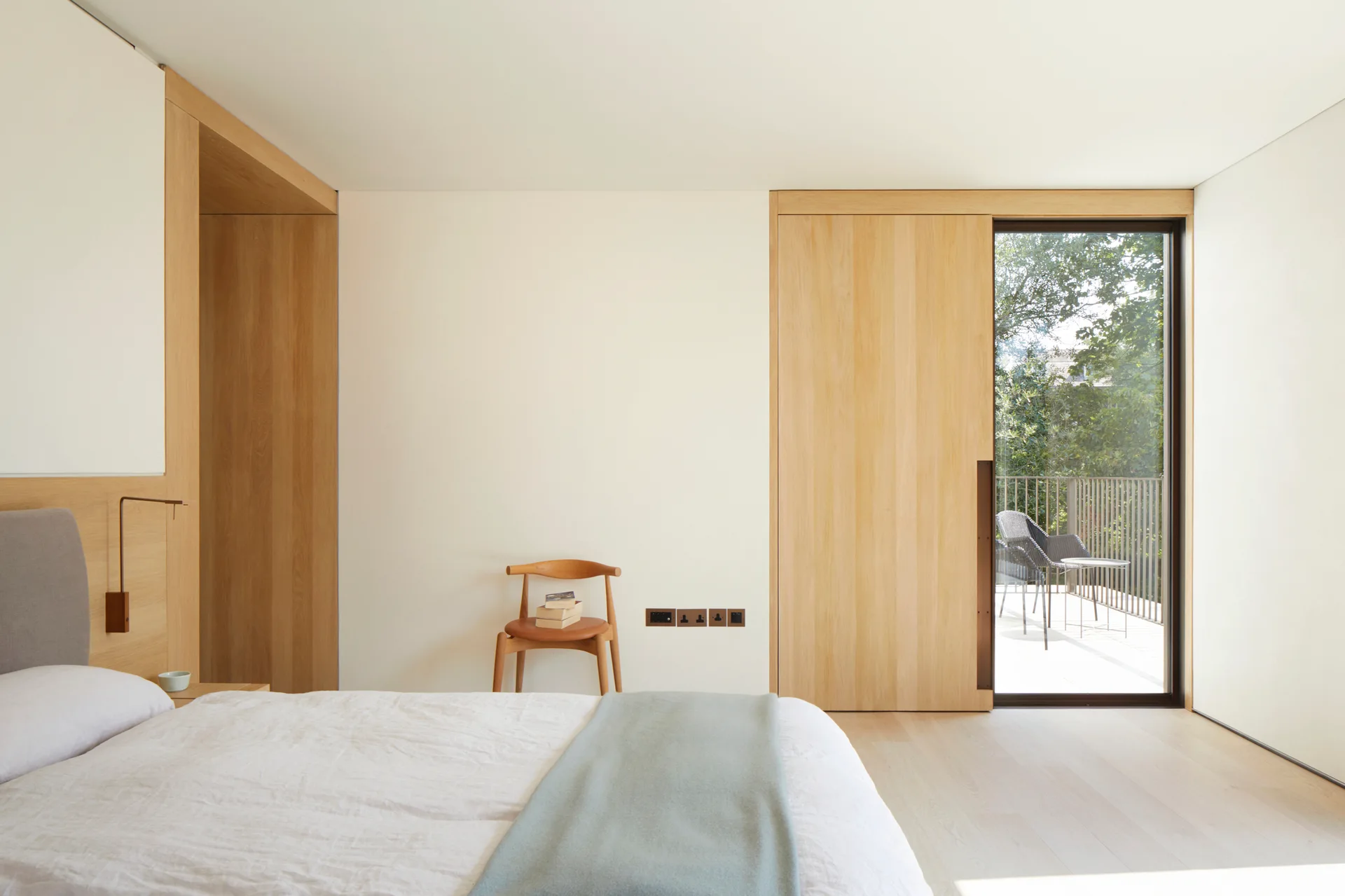 A minimalist bedroom featuring light wood wall panels, a low bed, a wooden accent chair, and a sliding door leading to an outdoor balcony.