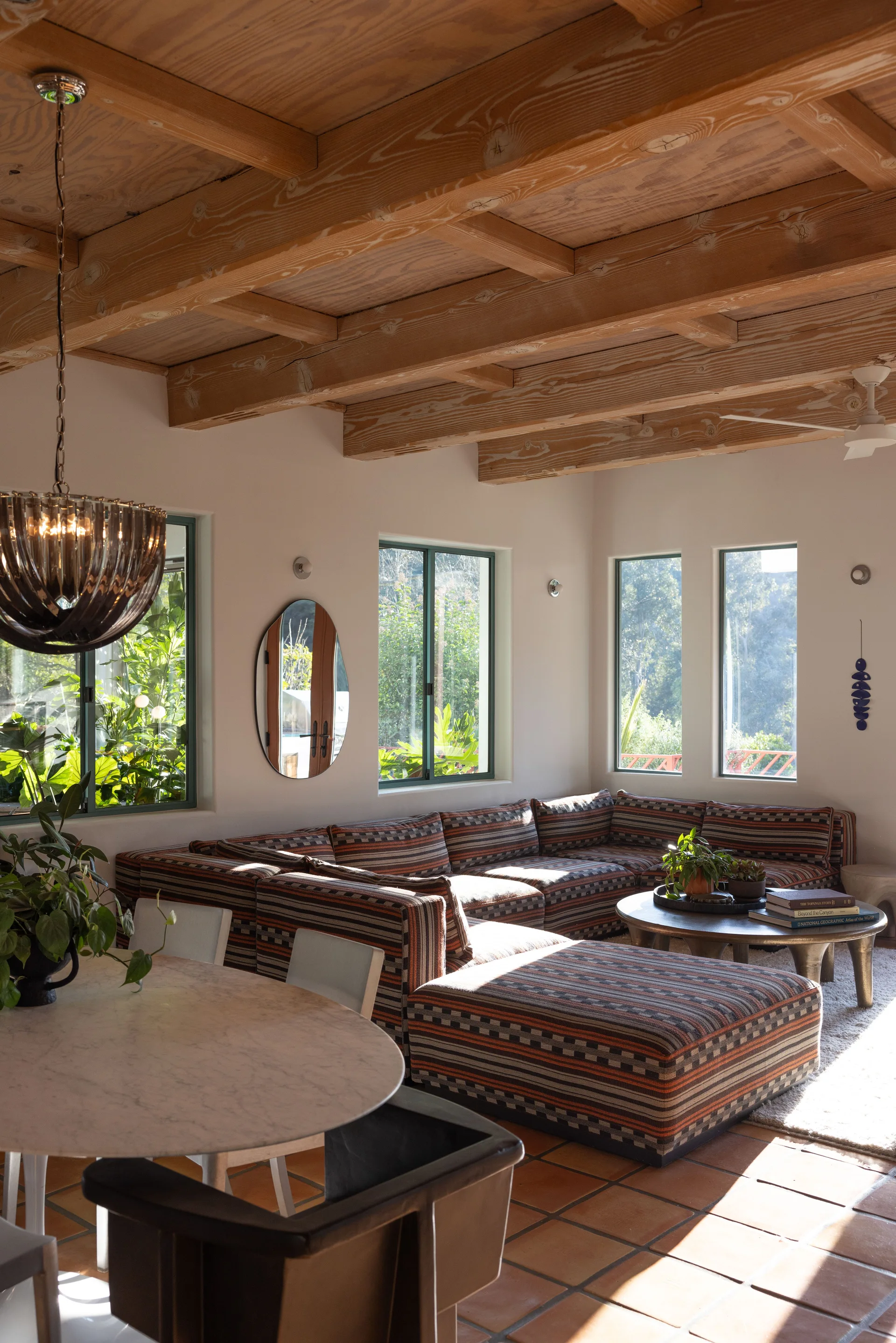 A sunlit living room featuring a patterned sectional sofa, terracotta tile floors, and an exposed wooden beam ceiling.