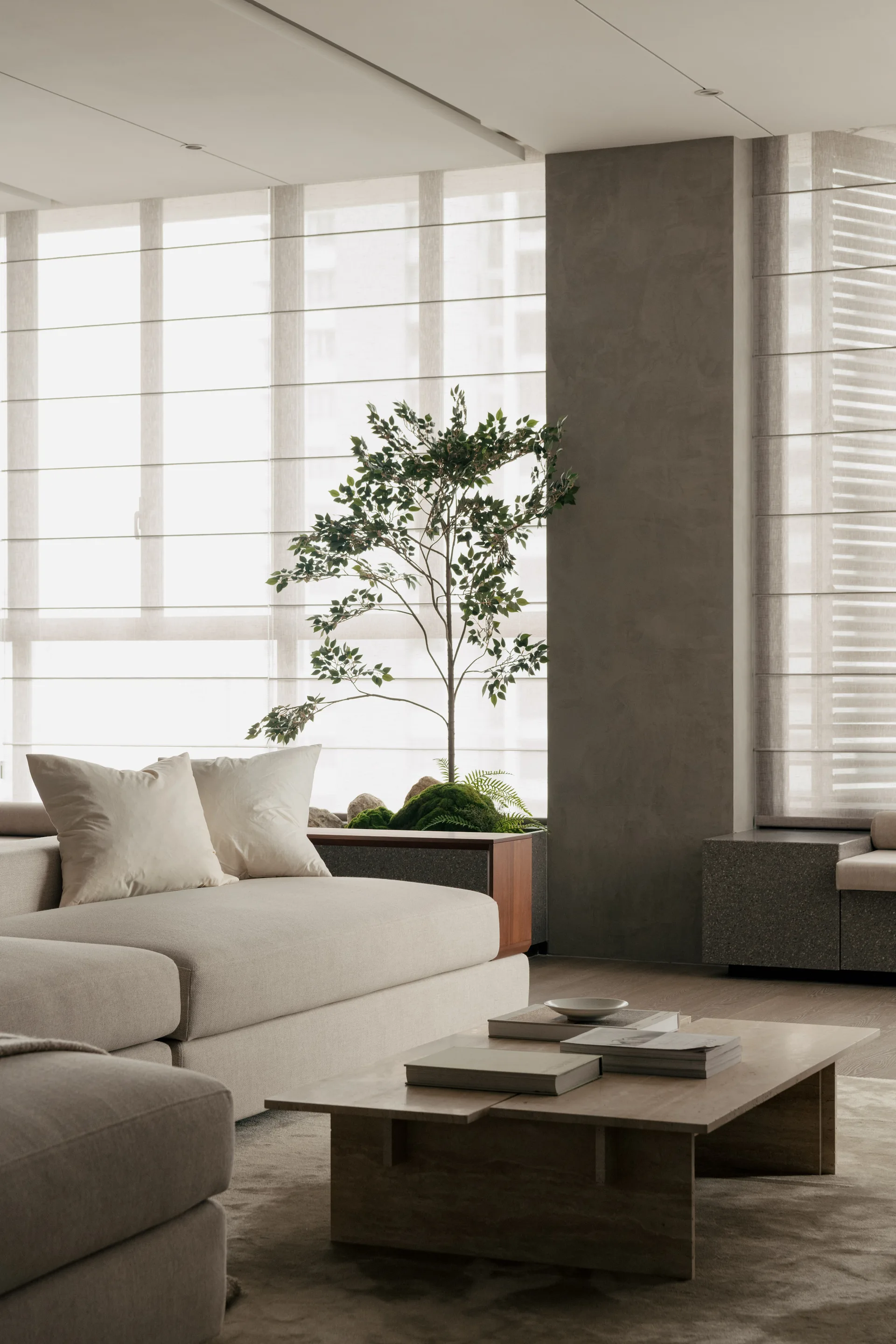 A minimalist living room with a cream upholstered sofa, a low wooden coffee table, and a small potted tree against translucent window blinds.