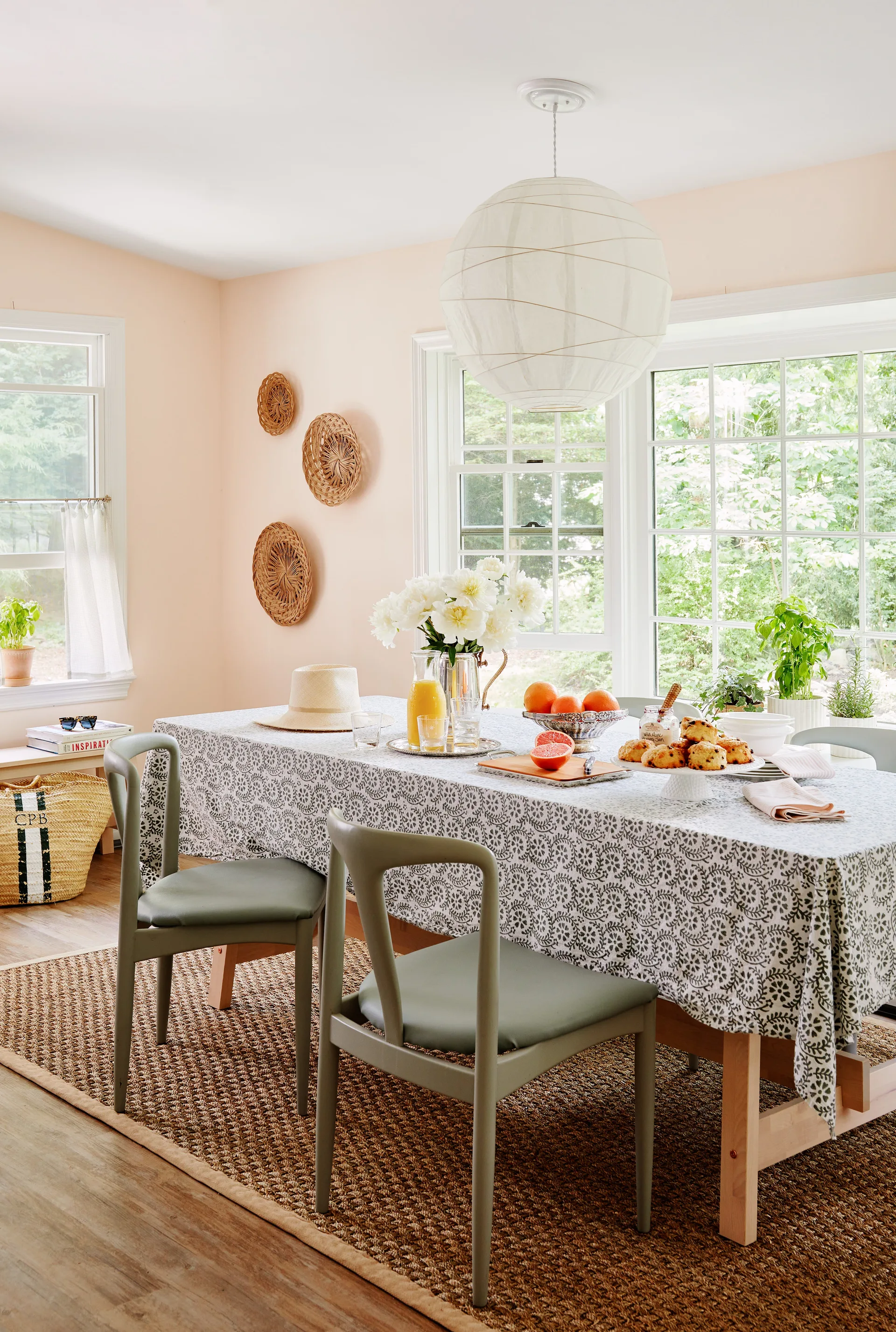A sunlit dining room features a long table with a floral tablecloth, olive green chairs, and a large paper lantern pendant under peach walls.