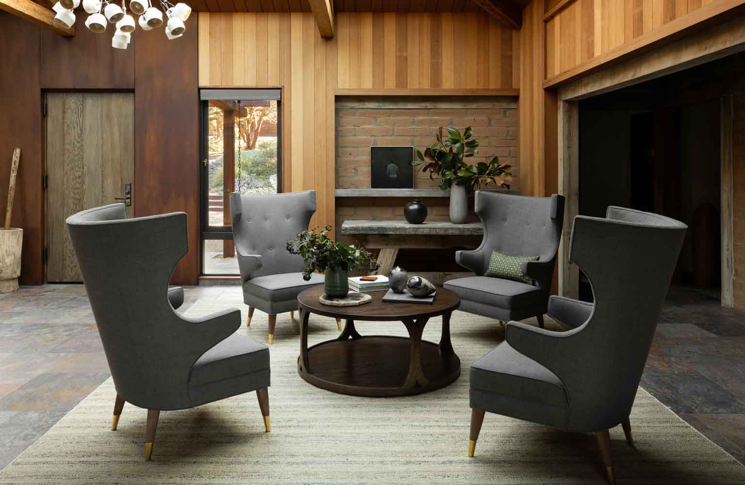 Four charcoal grey wingback armchairs are arranged around a round wood coffee table on a light-colored rug in a wood-paneled room.