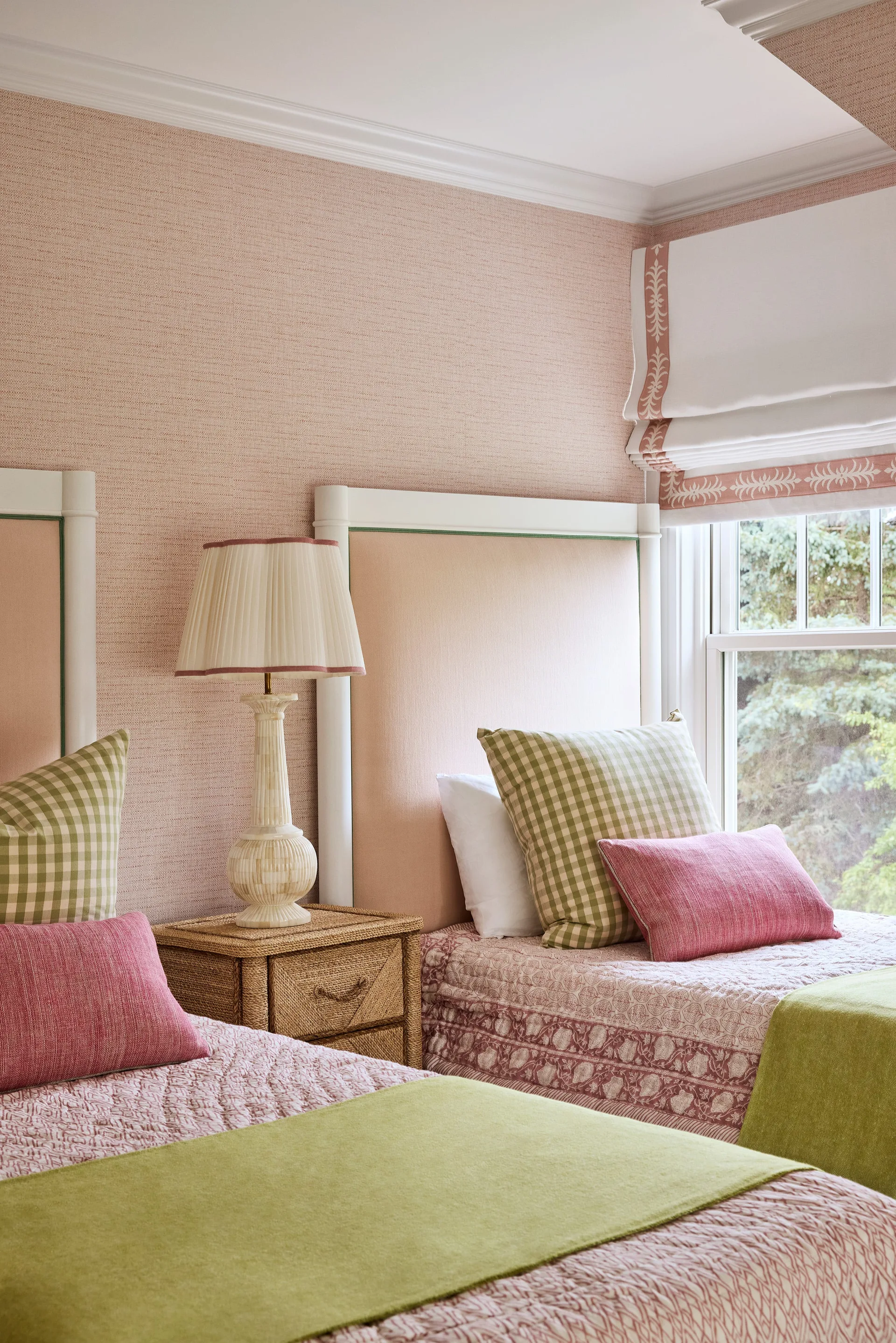 Two twin beds with pink upholstered headboards flank a woven wicker nightstand under a window with a bordered Roman shade.