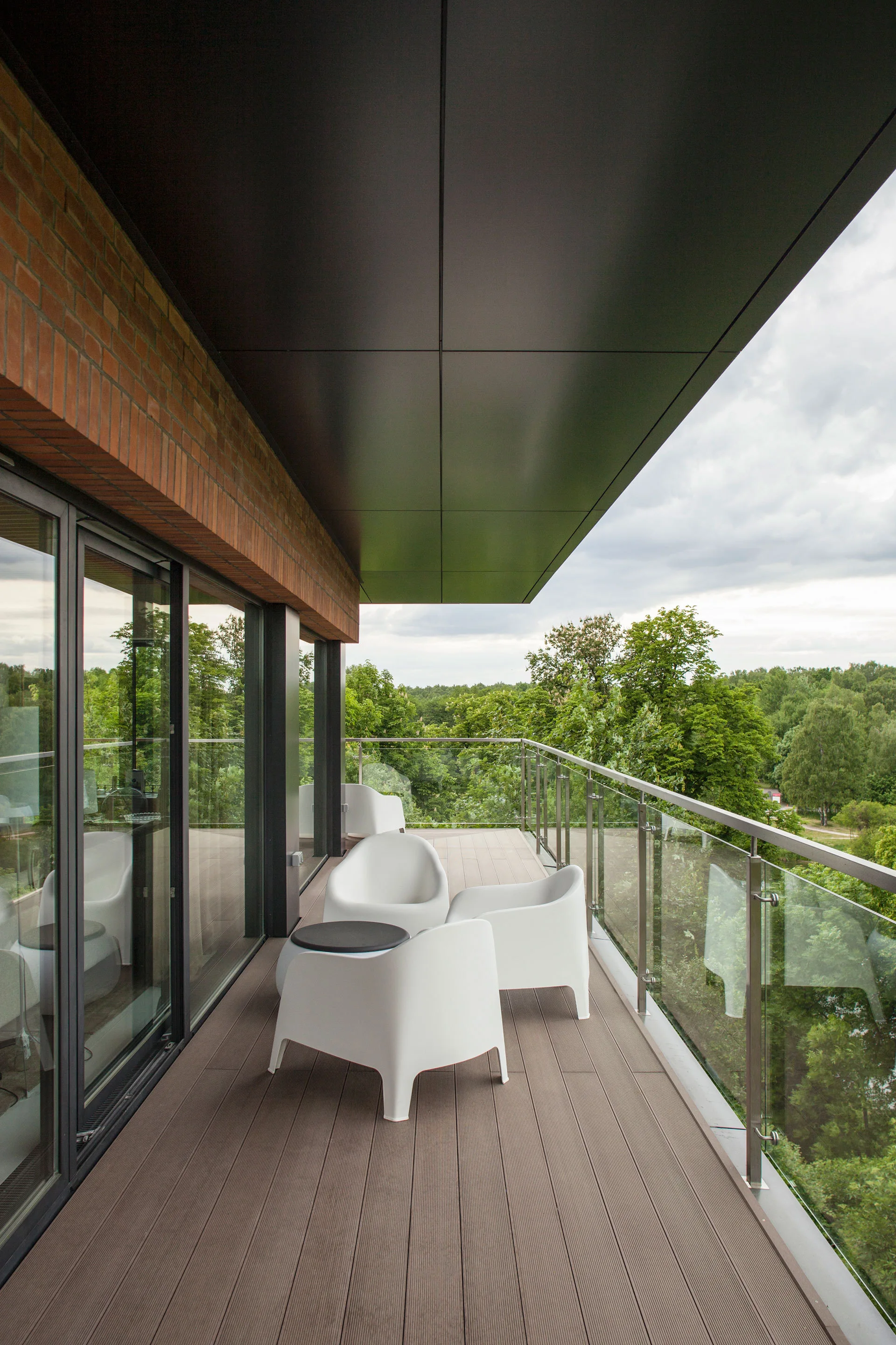 A long modern balcony features wood-look decking, curved white plastic armchairs, a glass railing, and large sliding glass doors.