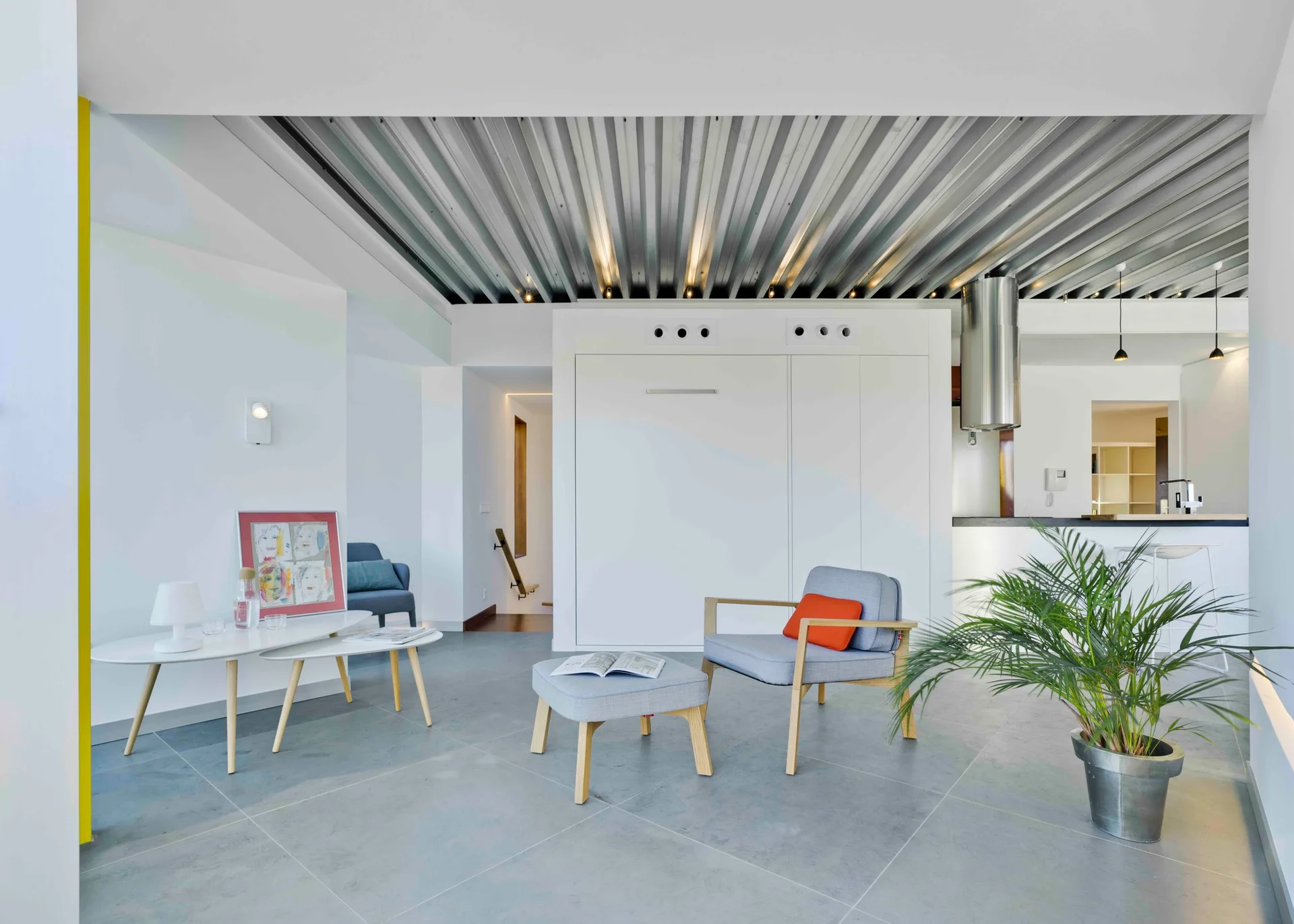 A minimalist living room with a corrugated metal ceiling, grey tiled floor, built-in white cabinets, and modern grey armchairs.