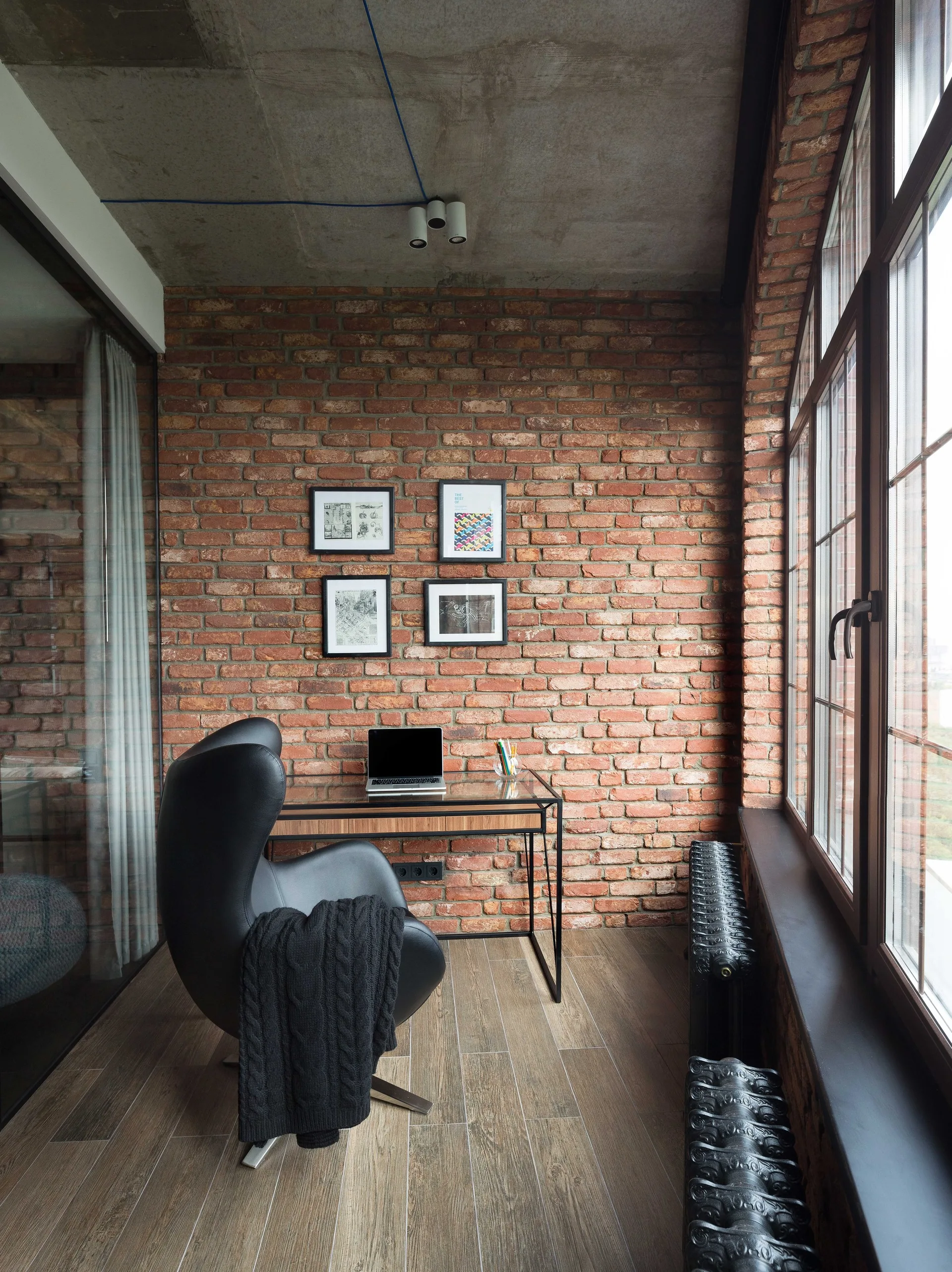 A modern industrial home office features an exposed brick wall, a raw concrete ceiling, a glass-topped desk with a laptop, and a curved black lounge chair.