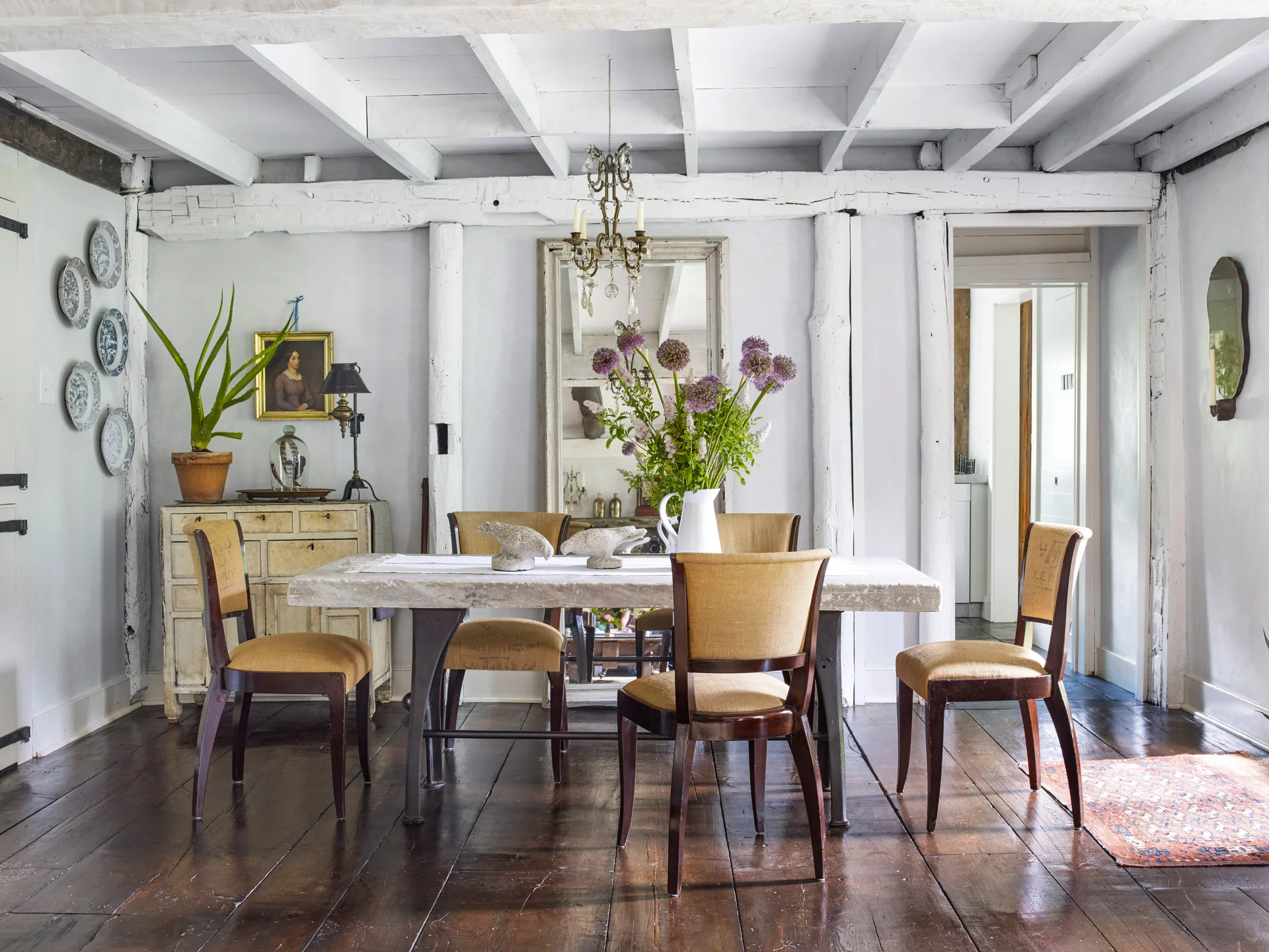 A rustic dining room features a stone-top table, upholstered chairs, white painted ceiling beams, and a large floor mirror behind the table.