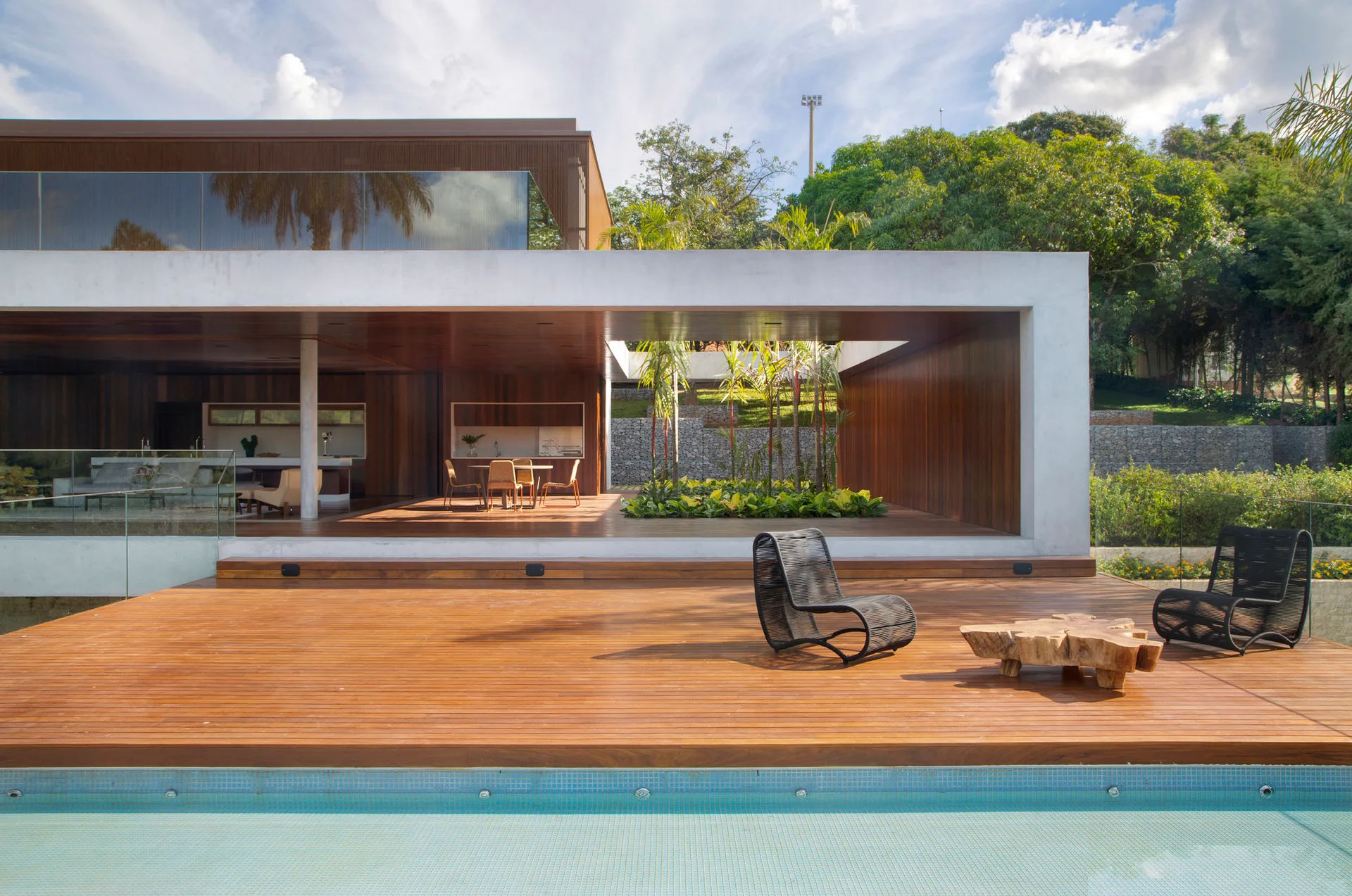 A modern concrete and wood home features a large wooden pool deck with black lounge chairs, a live-edge table, and an open covered terrace.