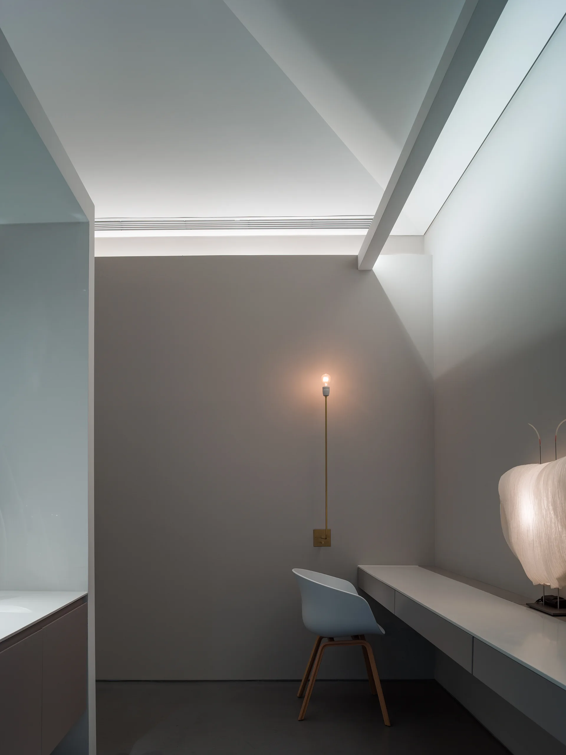 A minimalist home office features a white floating desk with a sculptural lamp, a modern gray shell chair, and a slender brass wall sconce.