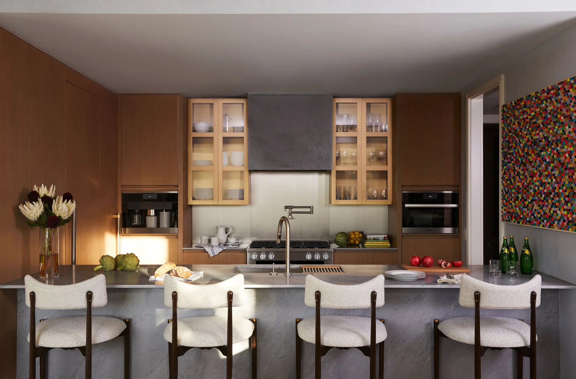A modern kitchen features a grey marble island with four upholstered bar stools, medium-toned wood cabinets, and glass-front upper storage.