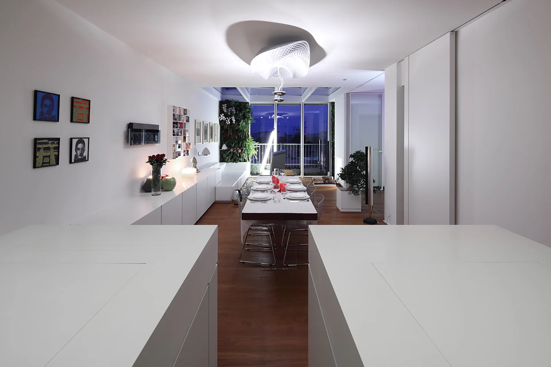A modern white dining room with a long dining table, a sculptural pendant light, sleek low sideboards, and a vertical green garden wall near the window.